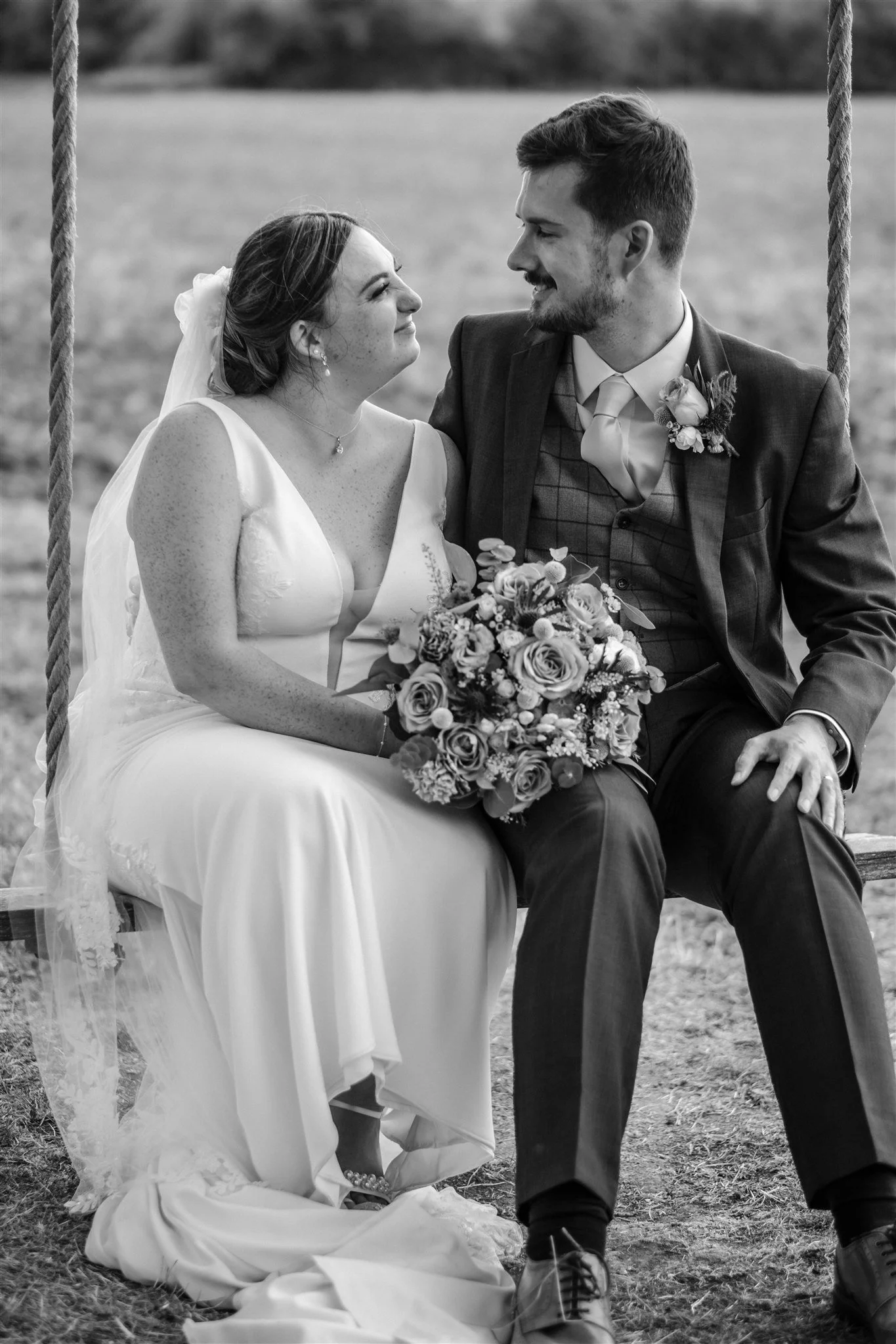 Black and white photo of a bride and groom sitting on a swing outdoors, facing each other and smiling. The bride wears a wedding dress, veil, and holds a bouquet. The groom wears a suit.