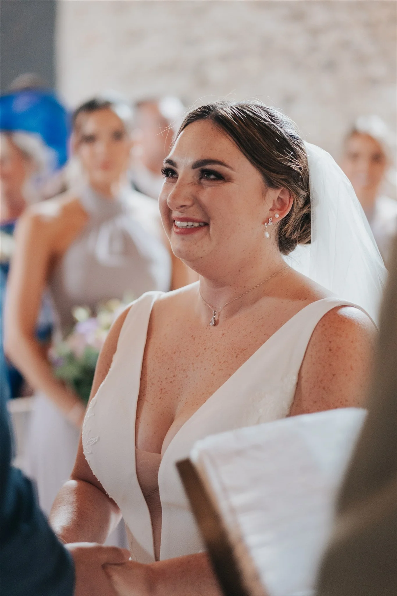 A bride smiling during her wedding ceremony, surrounded by blurred guests in the background.