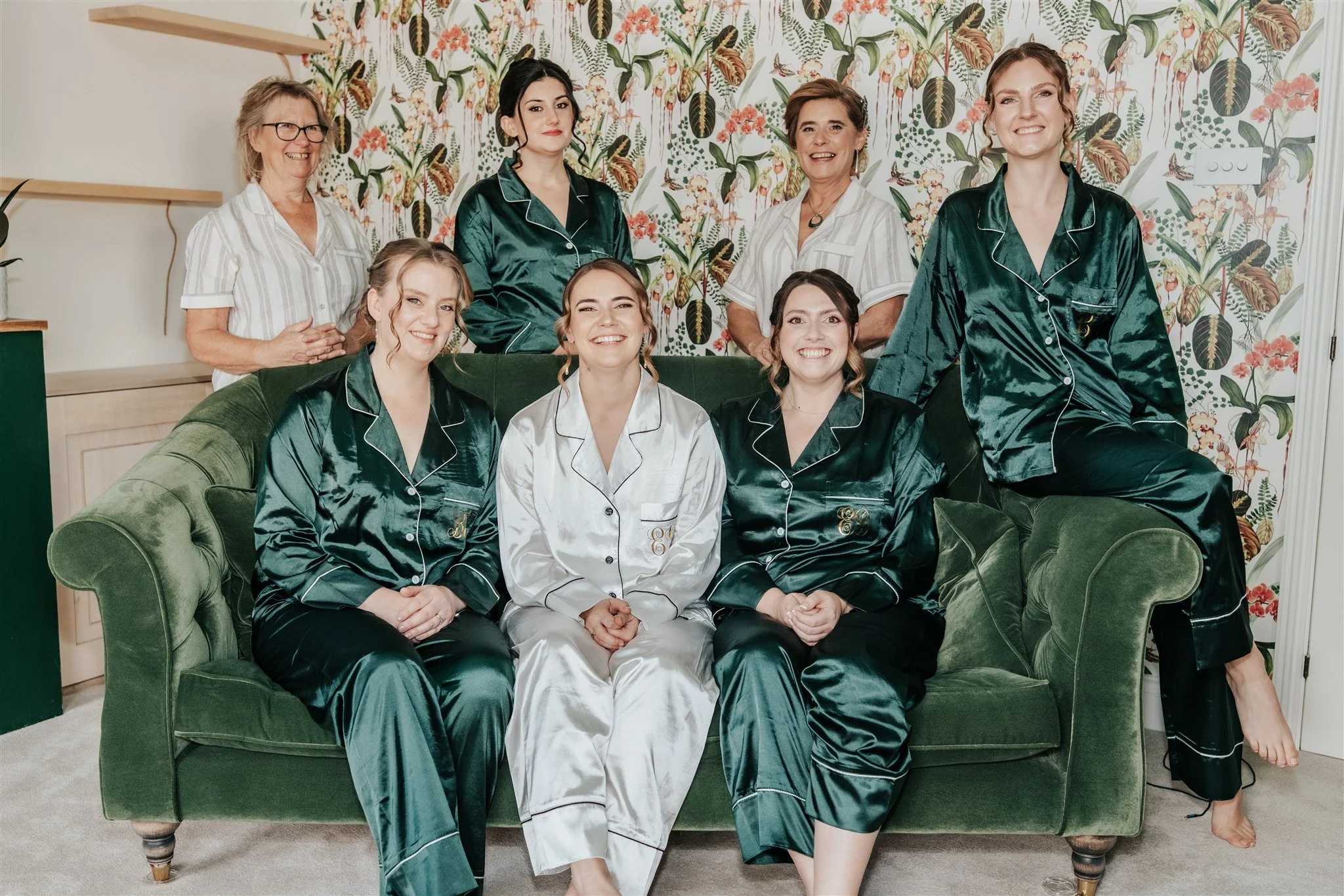 Group of women gathered in a living room, some seated on a green velvet sofa and others standing, smiling and wearing matching silky pajamas in green and white. The background features floral wallpaper with green, pink, and brown tones.
