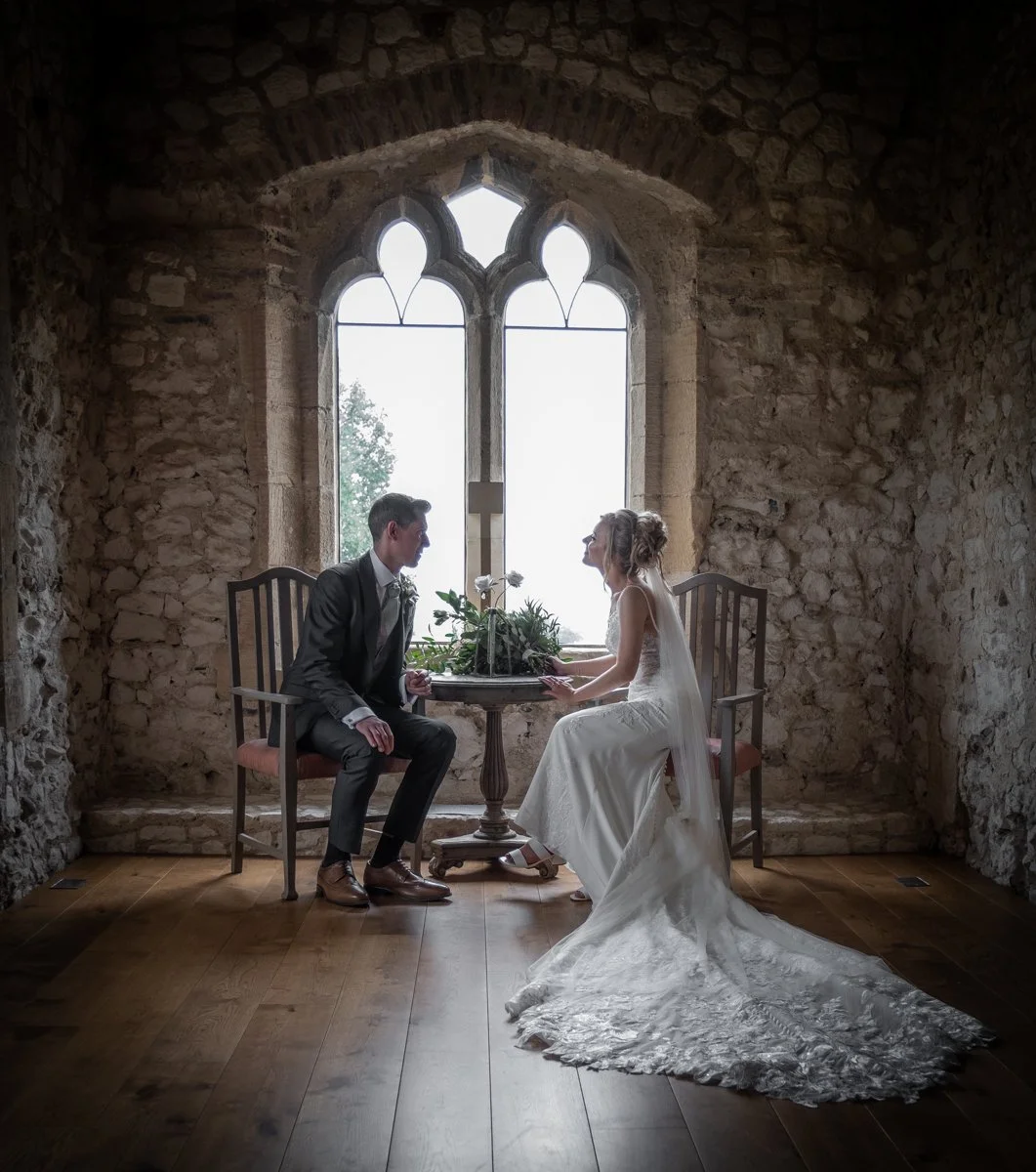 A bride and groom sitting across from each other at a small round table with flowers, in front of a large arched window with stone walls. The bride is in a white wedding dress with a train, and the groom is in a dark suit and tie.