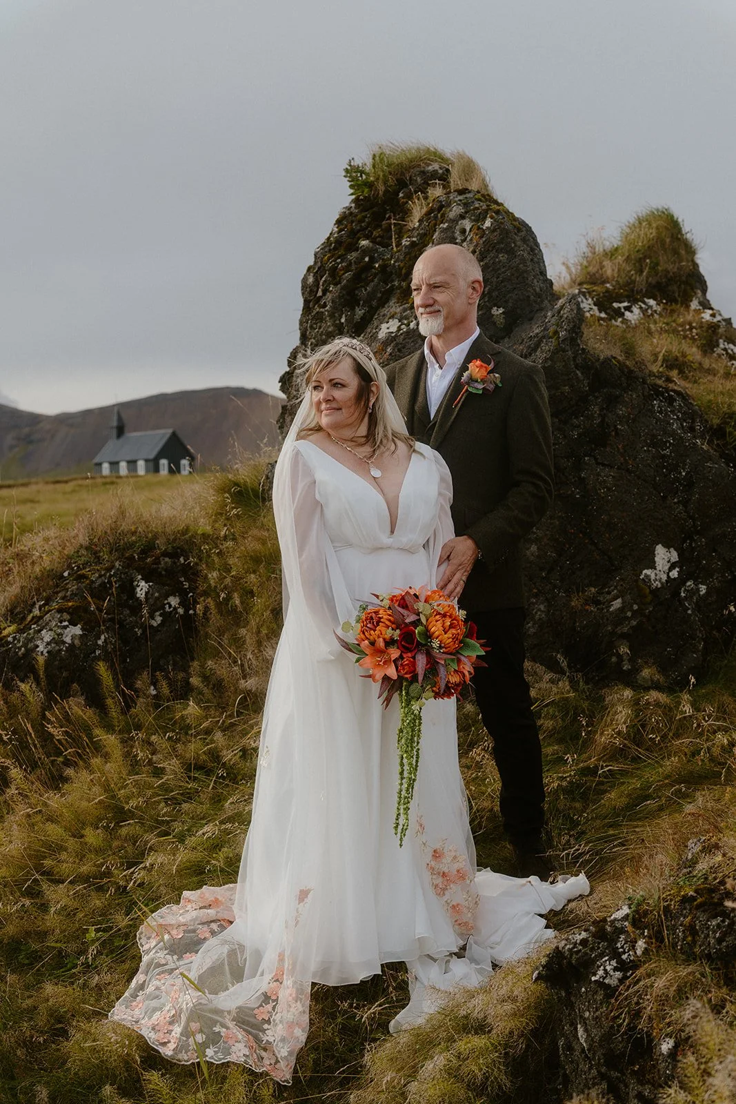A bride and a groom in wedding attire standing outdoors, with a small black church in the background, surrounded by grassy terrain and rocky formations.