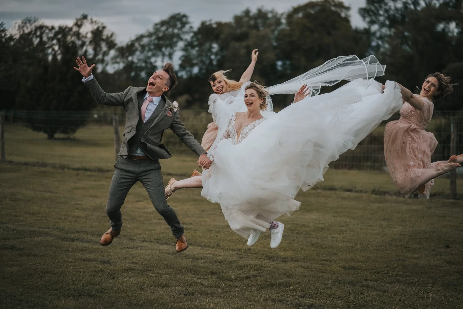 A group of people, including a bride in a white wedding dress and groom in a suit, jumping in the air outdoors on a grassy field during a wedding celebration, with a backdrop of trees and cloudy sky.