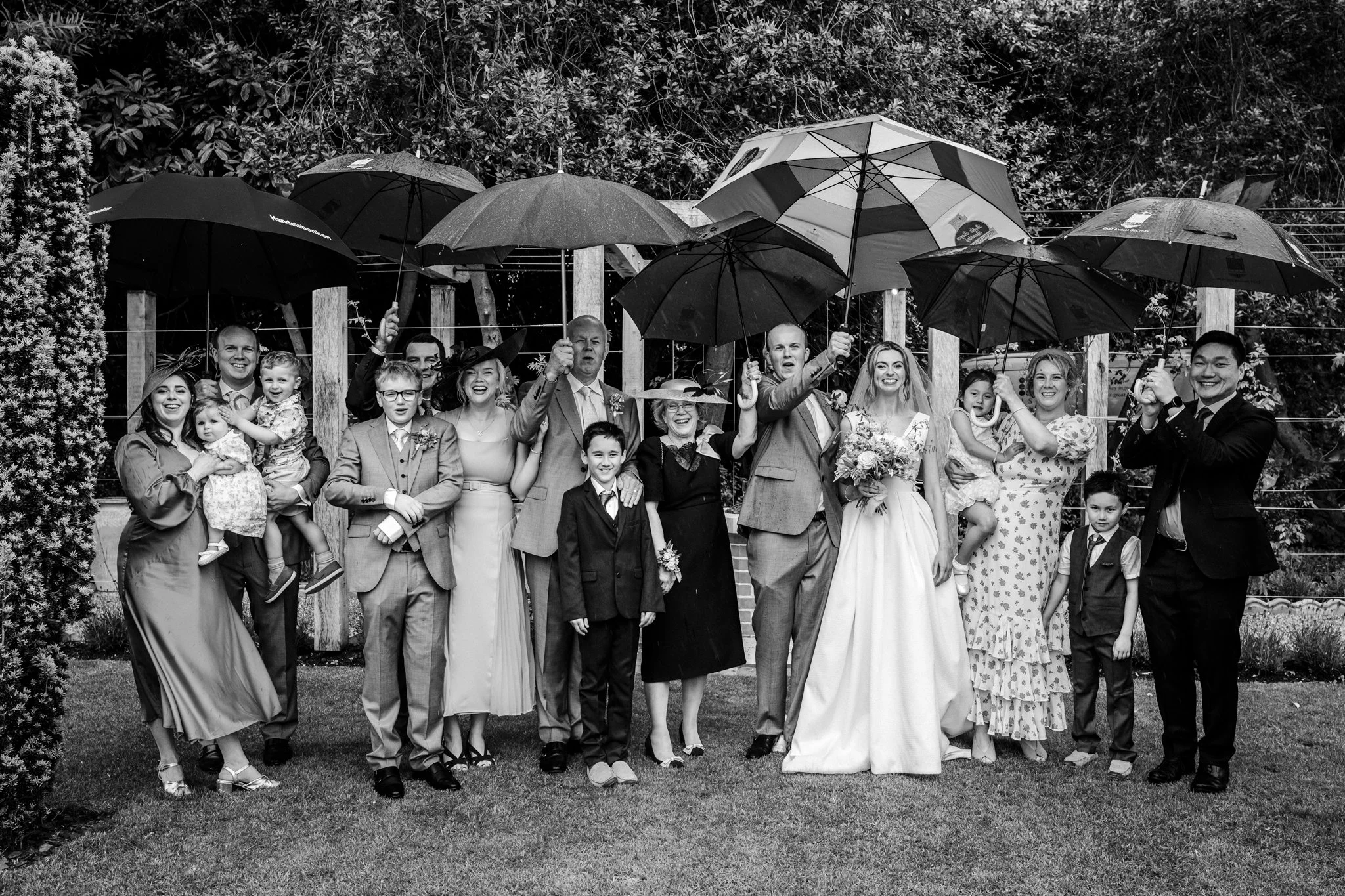 A group of people, including bride and groom, children and adults, dressed in formal wedding attire, standing outdoors in the rain holding umbrellas, smiling and celebrating.