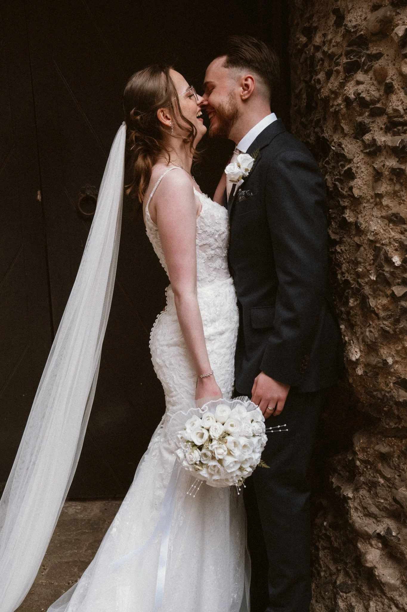 A bride and groom standing very close, smiling and touching noses, on their wedding day. The bride is dressed in a white lace wedding gown with a veil, holding a bouquet of white roses. The groom is wearing a dark suit with a white shirt and bow tie, and has a white flower boutonniere.