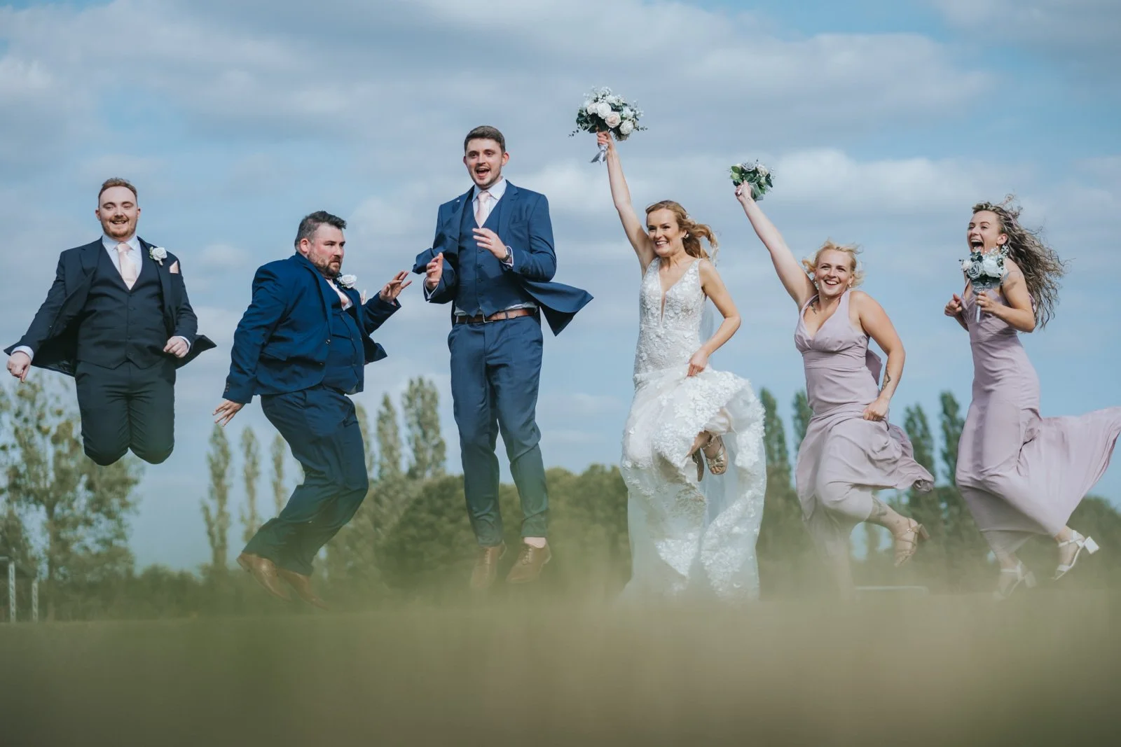 A group of wedding celebration people jumping in a field, with a blue sky and trees in the background. The bride is in a white wedding dress, holding a bouquet, and the groom along with four women in matching dresses are also jumping with excitement.