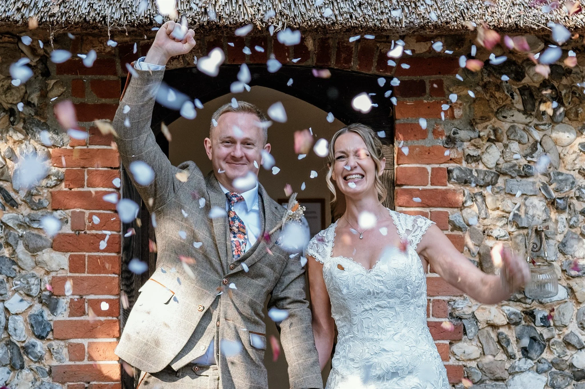 A newly married couple celebrating with confetti. The groom is wearing a check blazer and the bride is in a white lace wedding dress. They are standing under a brick archway.