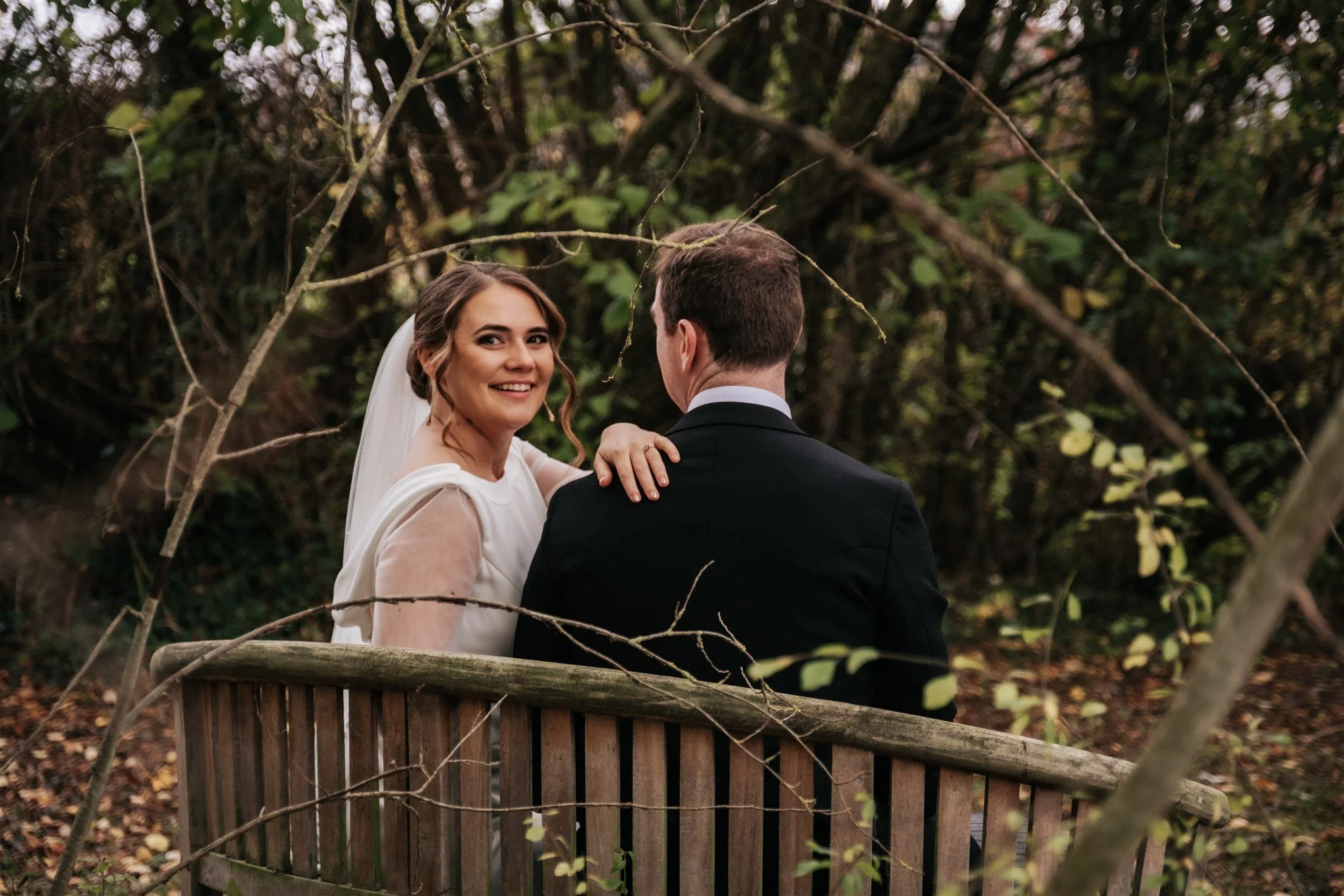 A bride and groom sharing a moment outdoors among trees and branches, with the bride smiling and looking at the camera. The bride is wearing a white dress and veil, and the groom is in a black suit, seen from behind.