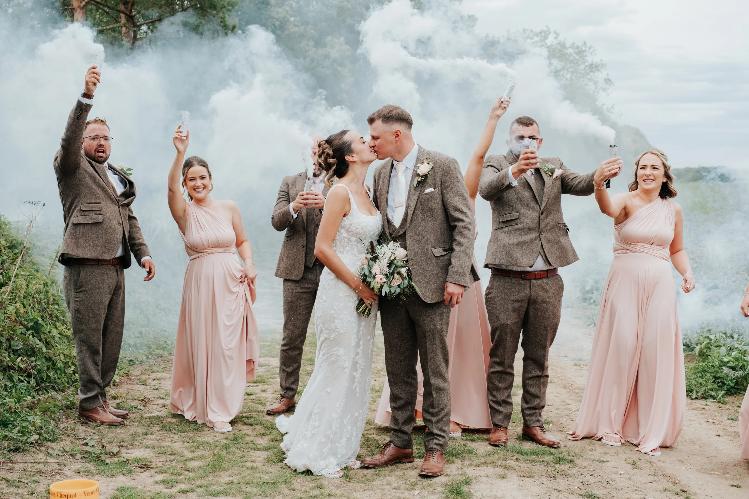 A wedding celebration outdoors with a bride and groom kissing, surrounded by wedding party members holding smoke bombs.