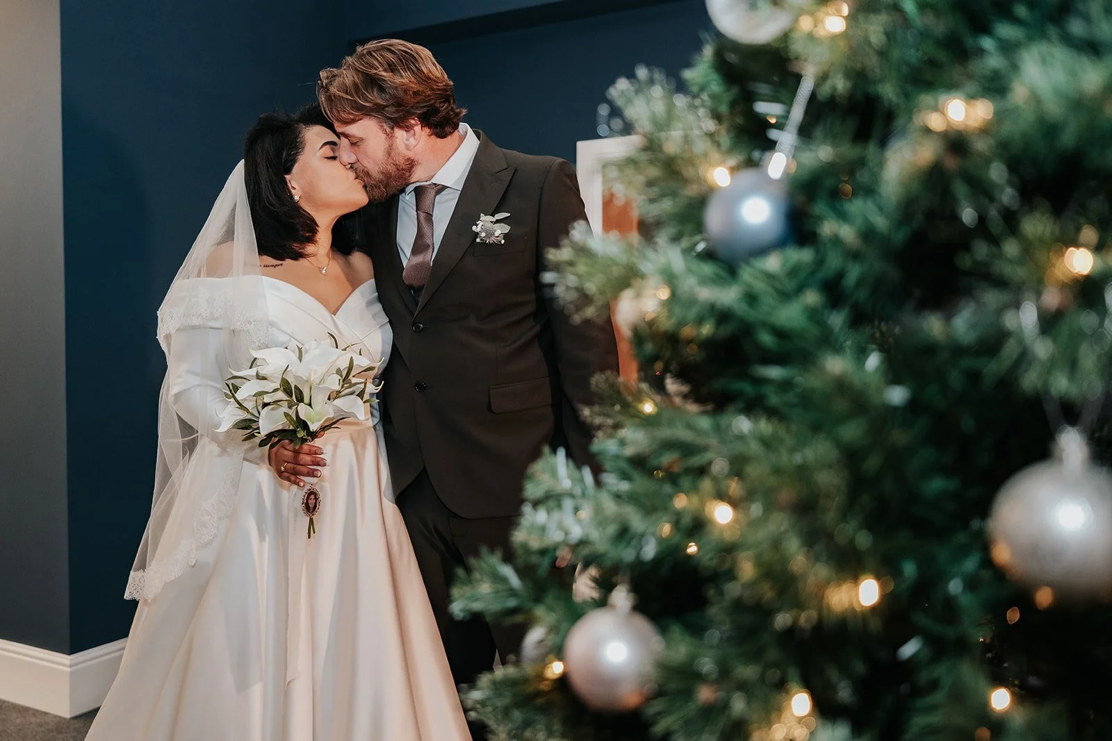 A bride and groom kissing at their wedding ceremony, with a decorated Christmas tree partially visible in the foreground.