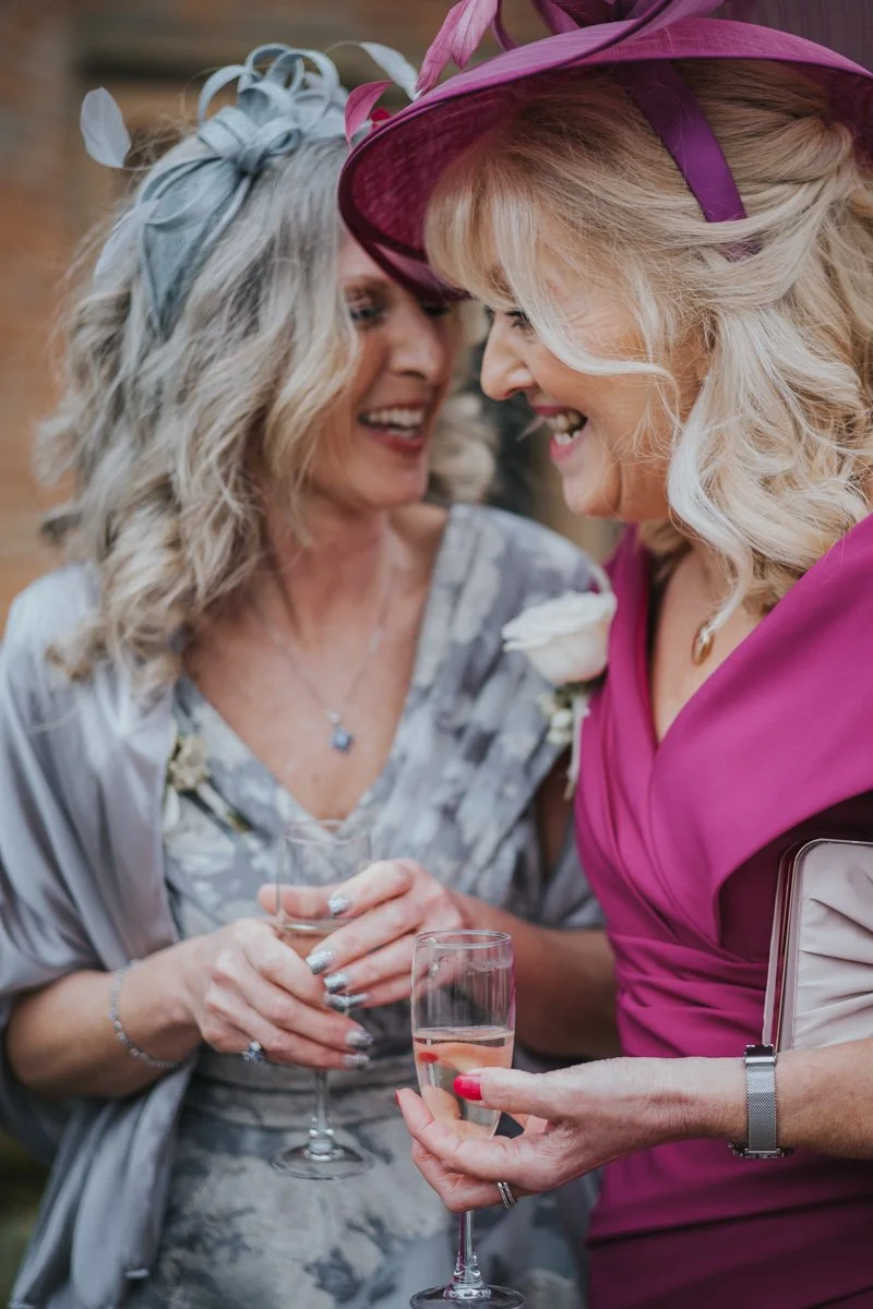 Two women sharing a joyful moment at a celebration, dressed elegantly, holding glasses of champagne, with wide smiles and close faces.