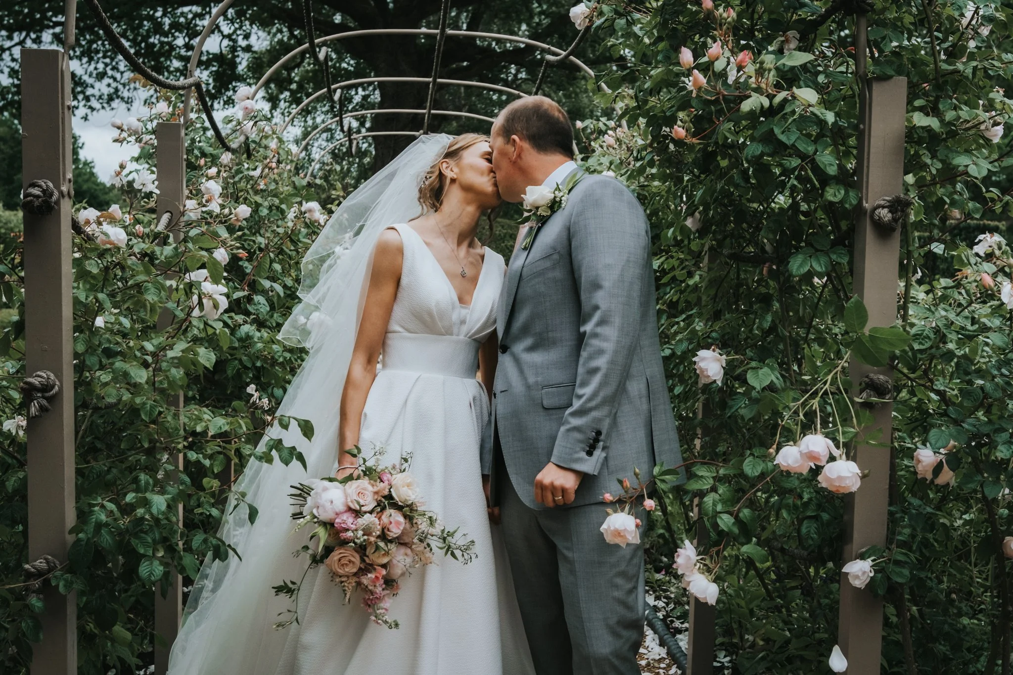 A bride and groom kissing outdoors amid greenery and pink and white flowers.