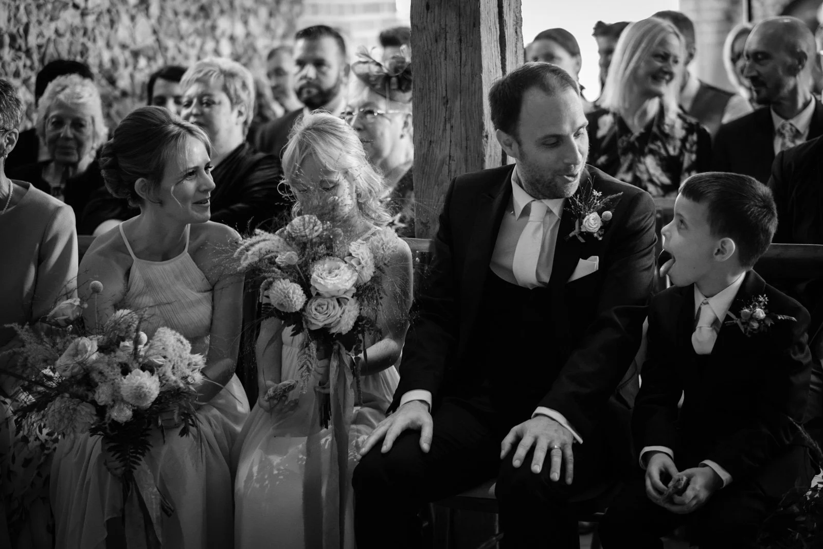 Black and white photo of a wedding ceremony with seated guests. The bride and groom are sitting next to each other, with the bride holding a bouquet and the groom making a playful face at a young boy sitting next to him. Guests in the background are 
