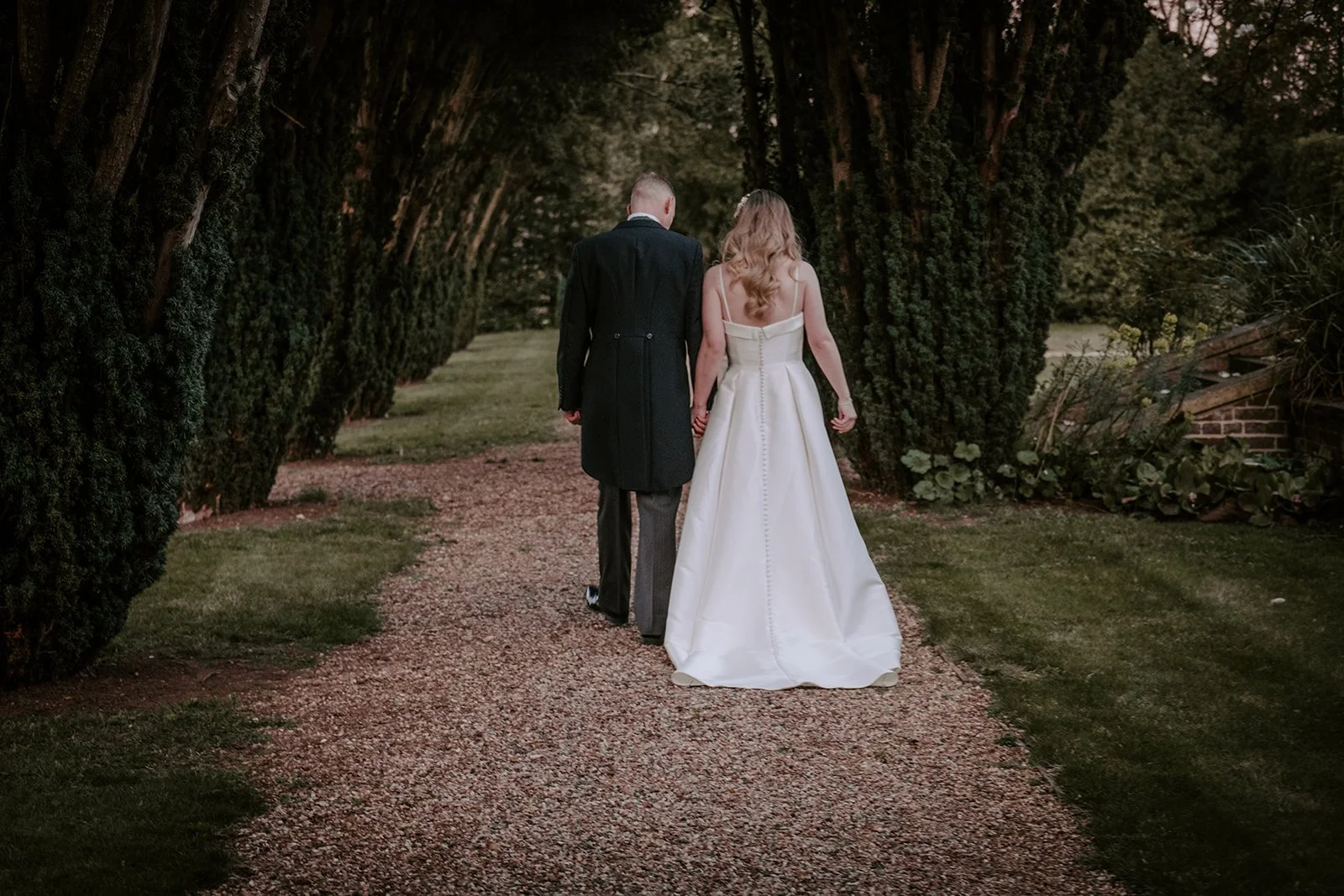 A bride and groom walking hand in hand on a gravel path lined with tall bushes, surrounded by lush greenery, during their wedding at Lanwades Hall, Newmarket, Suffolk.