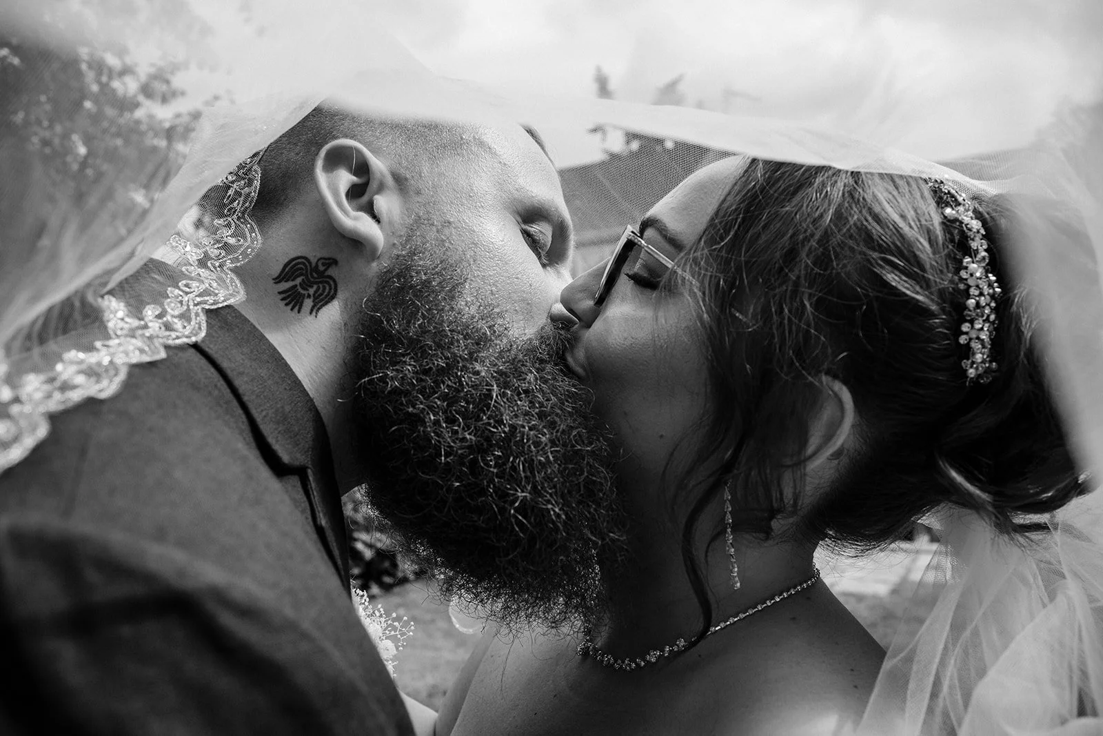 A black-and-white photo of a newlywed couple kissing, with the groom wearing a veil and a tattoo on his neck, and the bride wearing glasses, a tiara, and a veil.