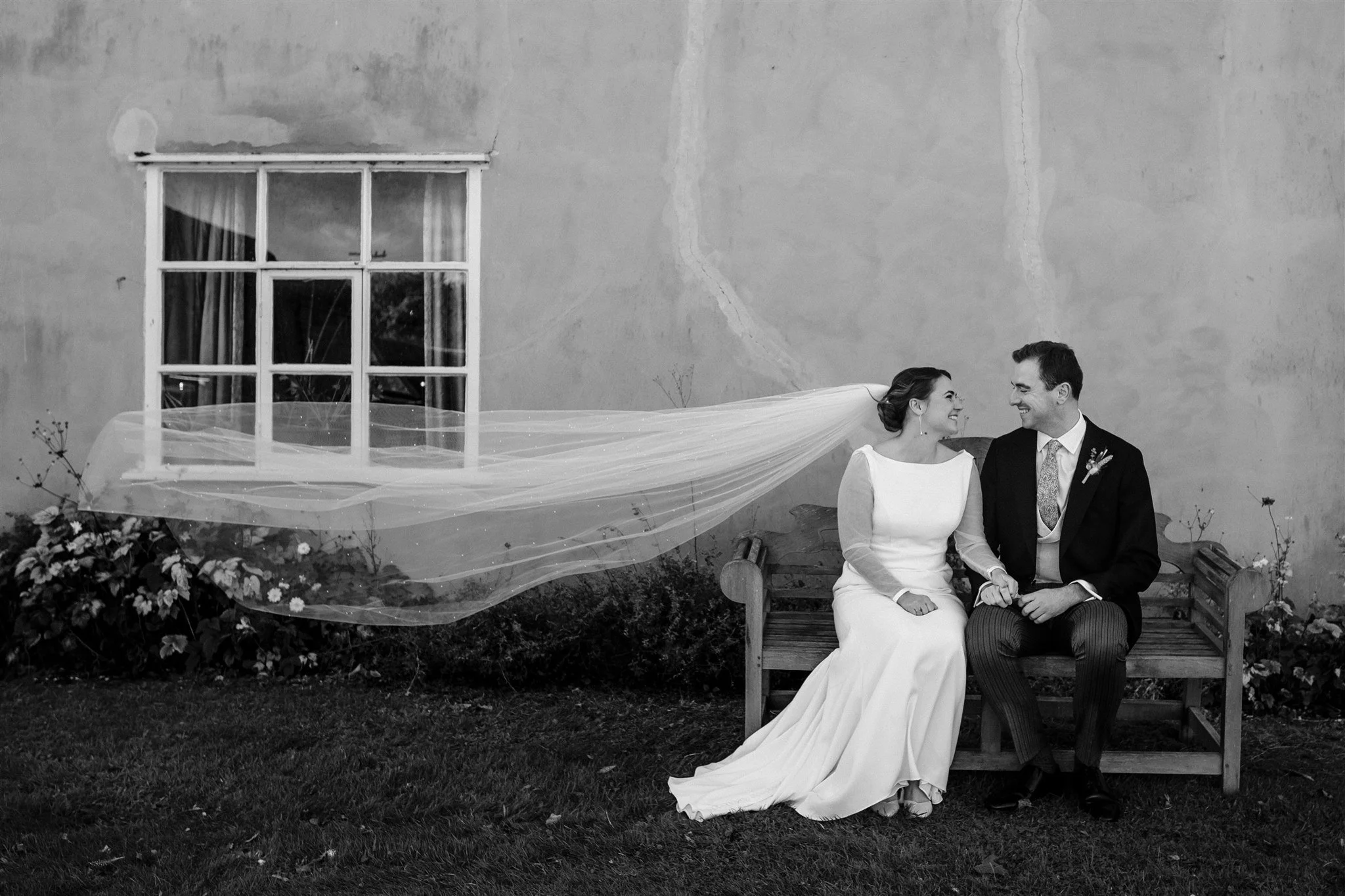 A bride and groom sitting on a wooden bench outdoors, holding hands and smiling at each other, with a large, textured wall, a window, and plants in the background at Alpheton Hall Barns, Suffolk.