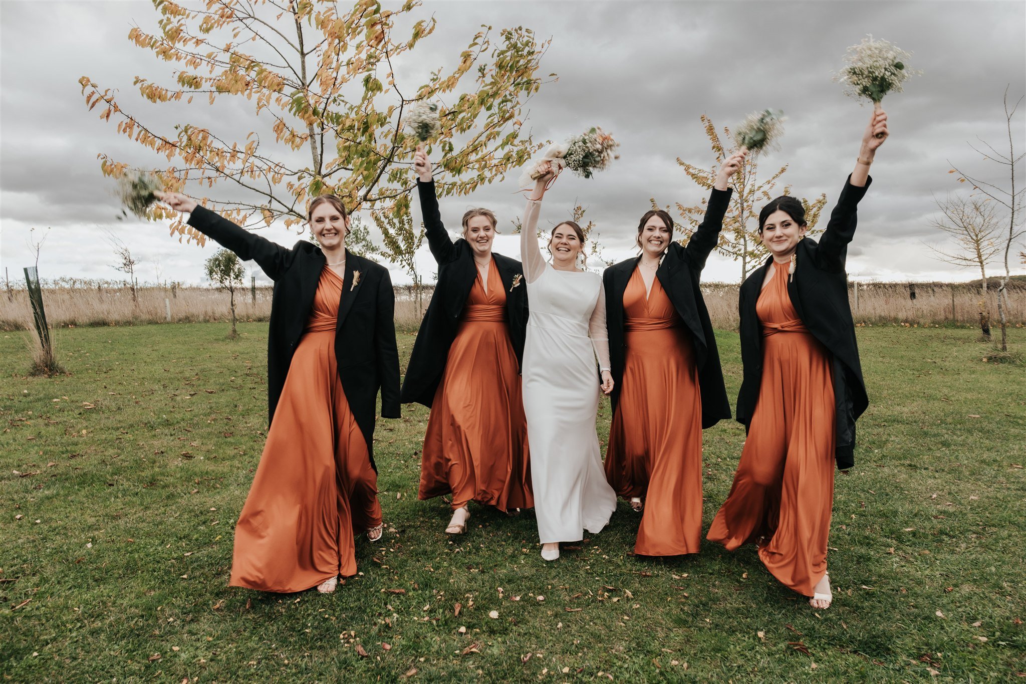 Bride and bridesmaids wearing suit jackets celebrating at Alpheton Hall Barns wedding venue, holding bouquets of flowers full of baby's breath, with a tree and cloudy sky in the background.