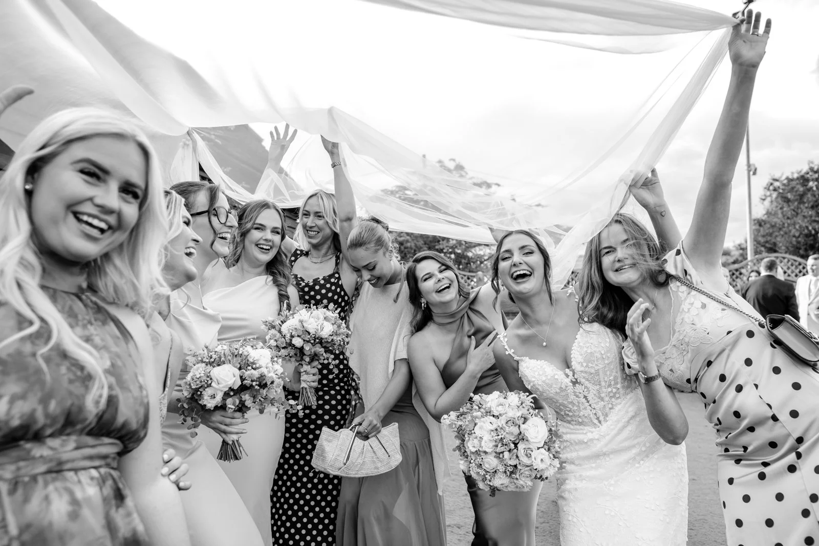A group of women at a celebration, smiling and holding bouquets of flowers, standing under a canopy canopy outdoors.