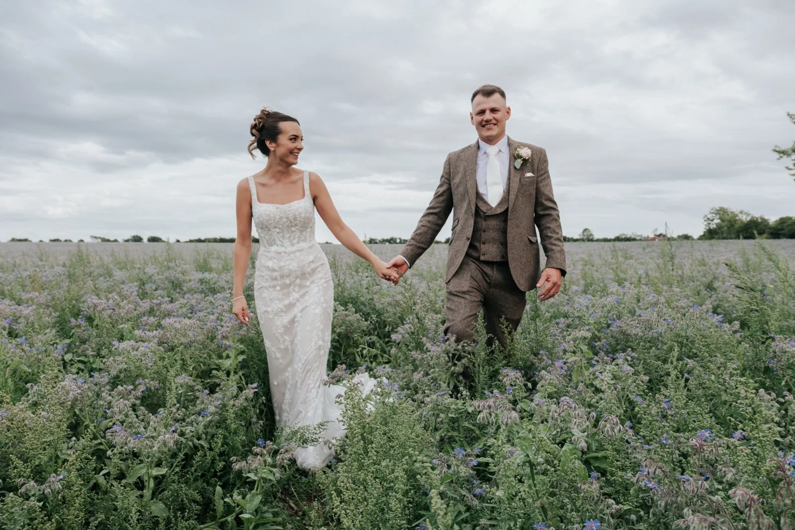 A bride and groom walking hand-in-hand through a field of purple wildflowers on a cloudy day, smiling and looking at each other.