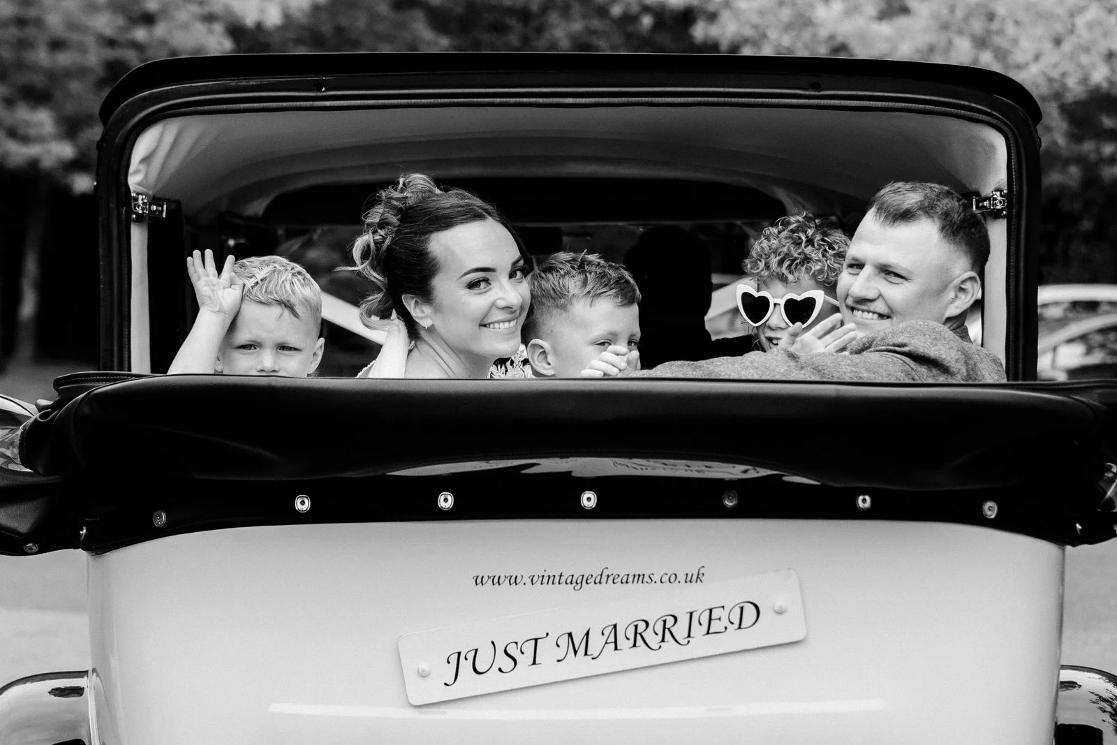 A black and white photo of a smiling bride and groom with their children, riding in a vintage car with a "Just Married" sign, celebrating their wedding.