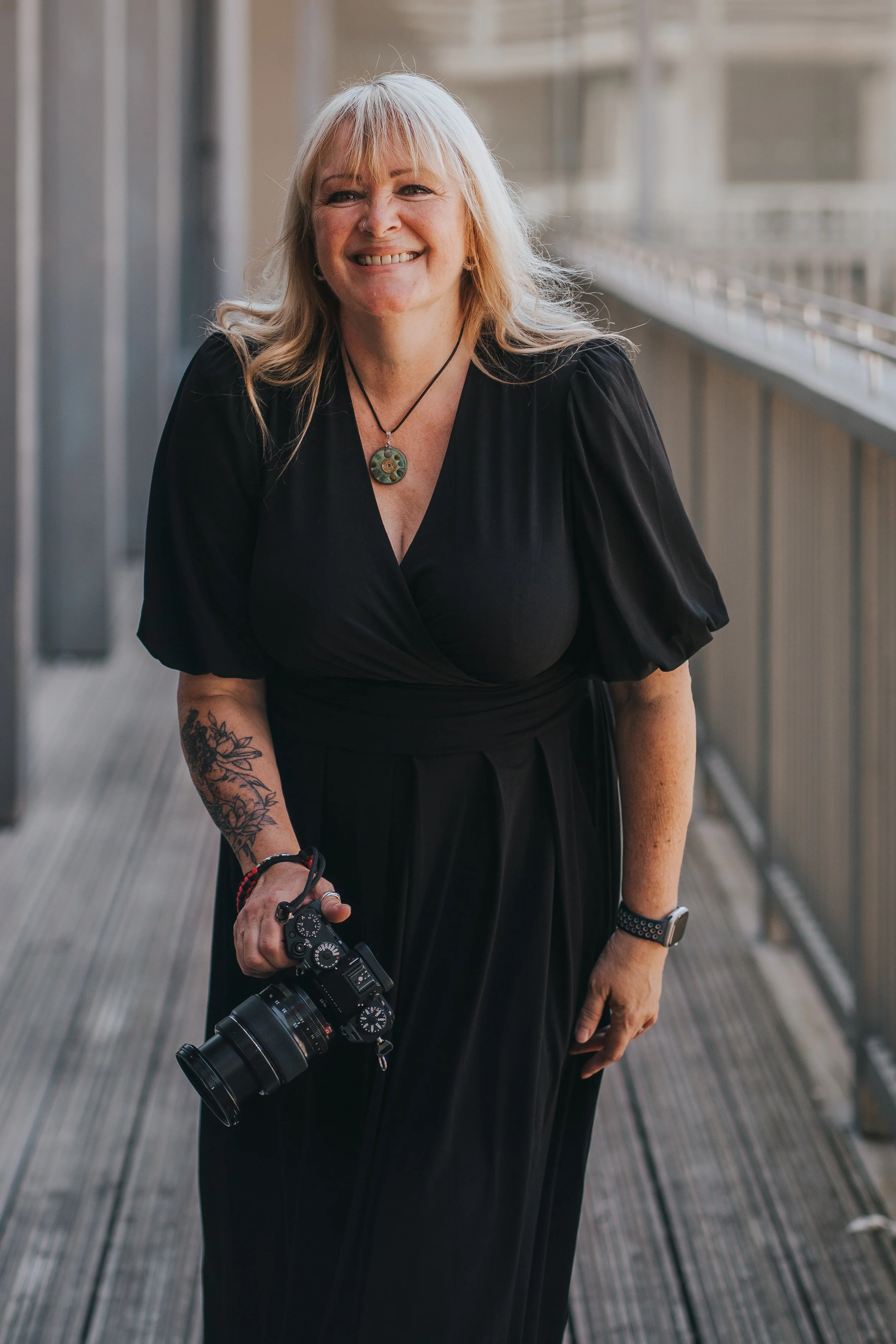 Jackie Minns holding a professional camera, standing on a wooden walkway near a building with balconies.