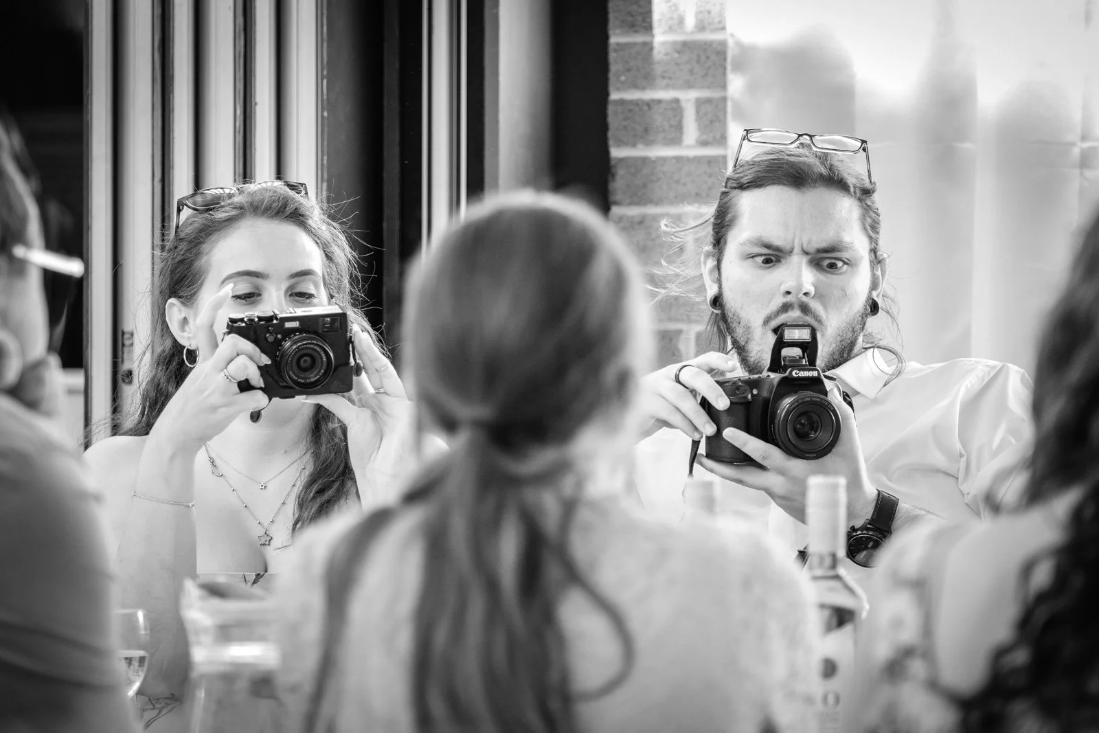 Black and white candid photo of people taking pictures with cameras at a wedding, with a woman in the background looking at her camera and a man in the foreground reacting to his camera.
