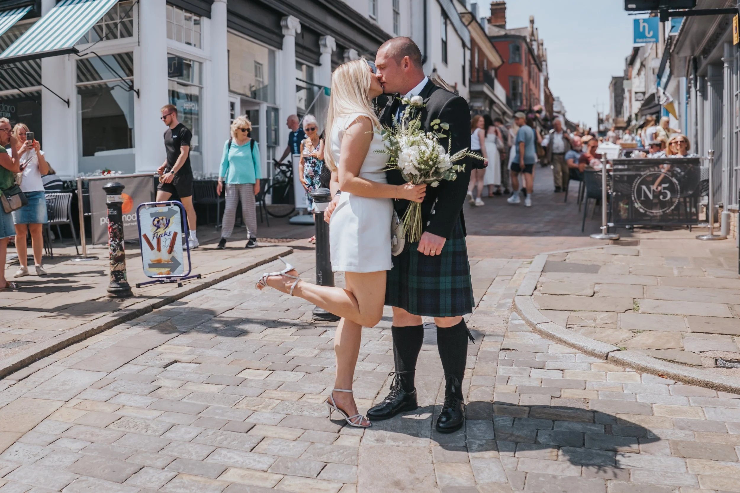 A newlywed couple sharing a kiss at the bottom of Abbeygate Street, on Angel Hill Bury St Edmunds with onlookers and shops in the background.