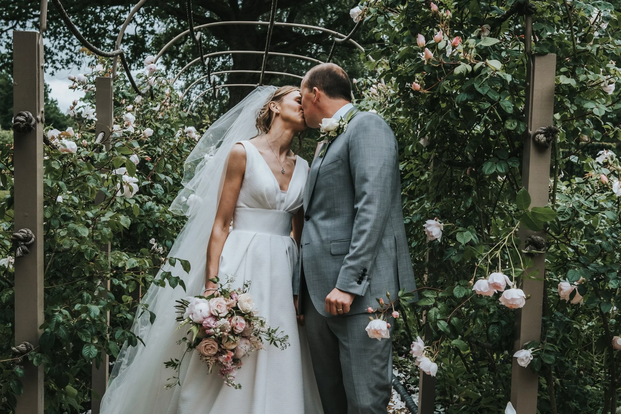 A bride and groom in Suffolk, sharing a kiss outdoors during their wedding, surrounded by greenery and flowering bushes.