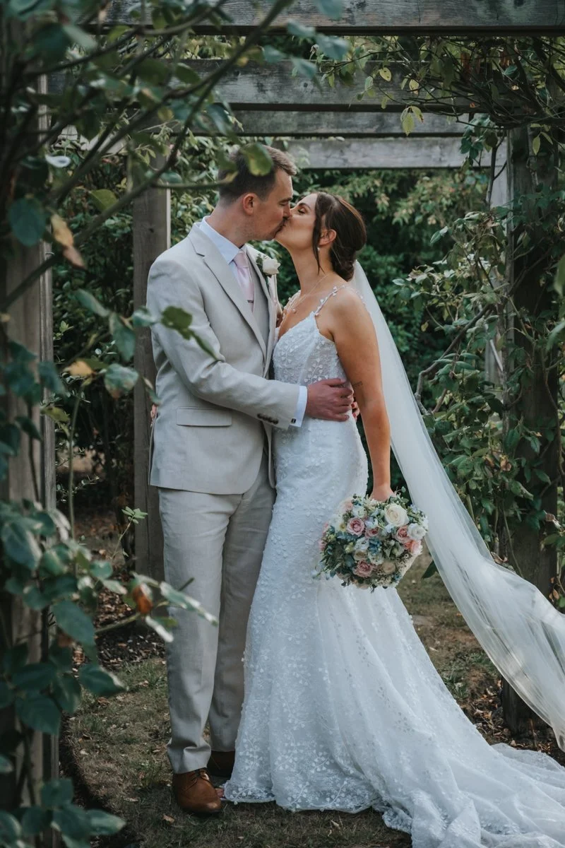A bride and groom sharing a kiss during their wedding in an outdoor garden setting, with green foliage and a rustic wooden arch in the background.