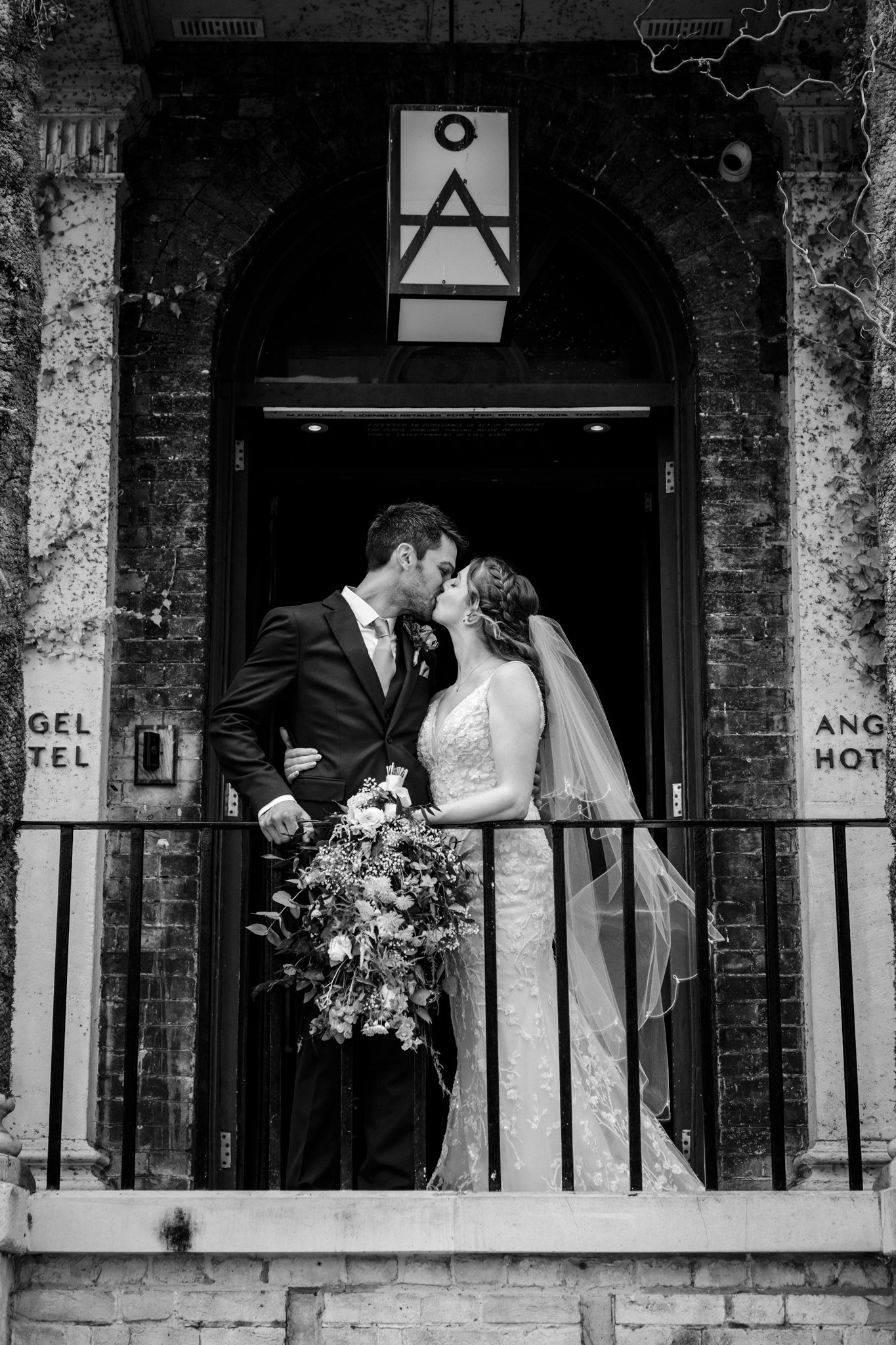 Black and white photo of a bride and groom kissing on a balcony at the Angel Hotel Bury St Edmunds . The groom is in a tuxedo, and the bride is in a lace wedding dress with a veil.