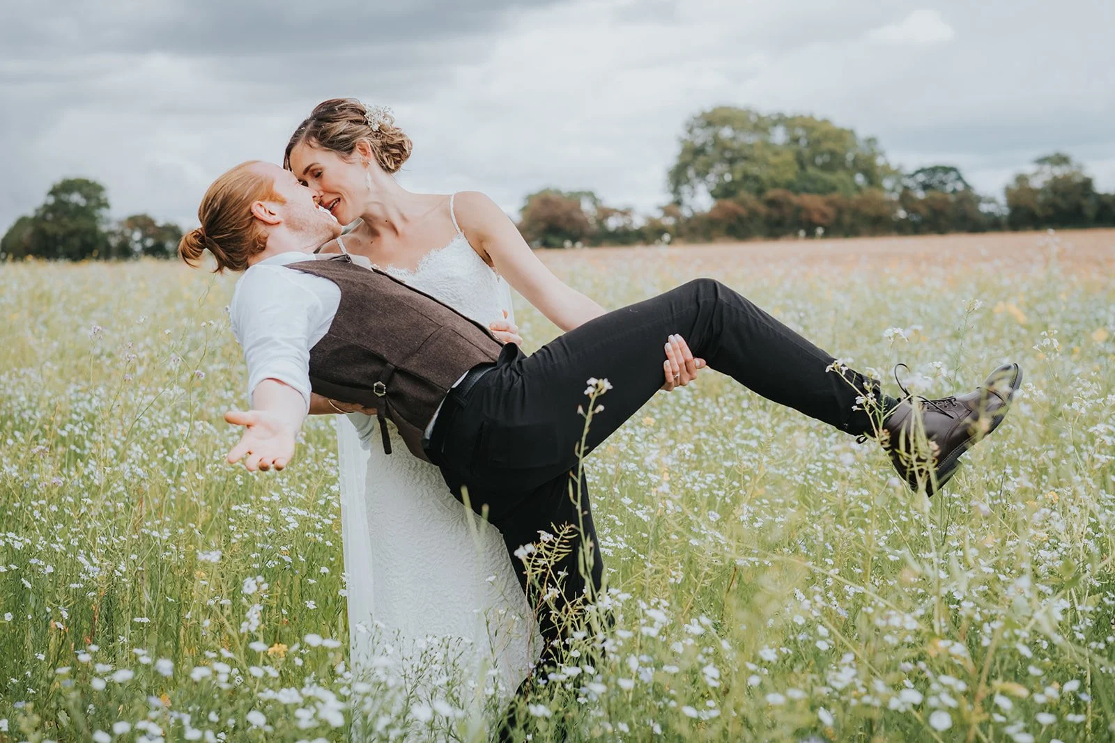 A newlywed couple in wedding attire enjoying a moment in a field of wildflowers with trees in the background.