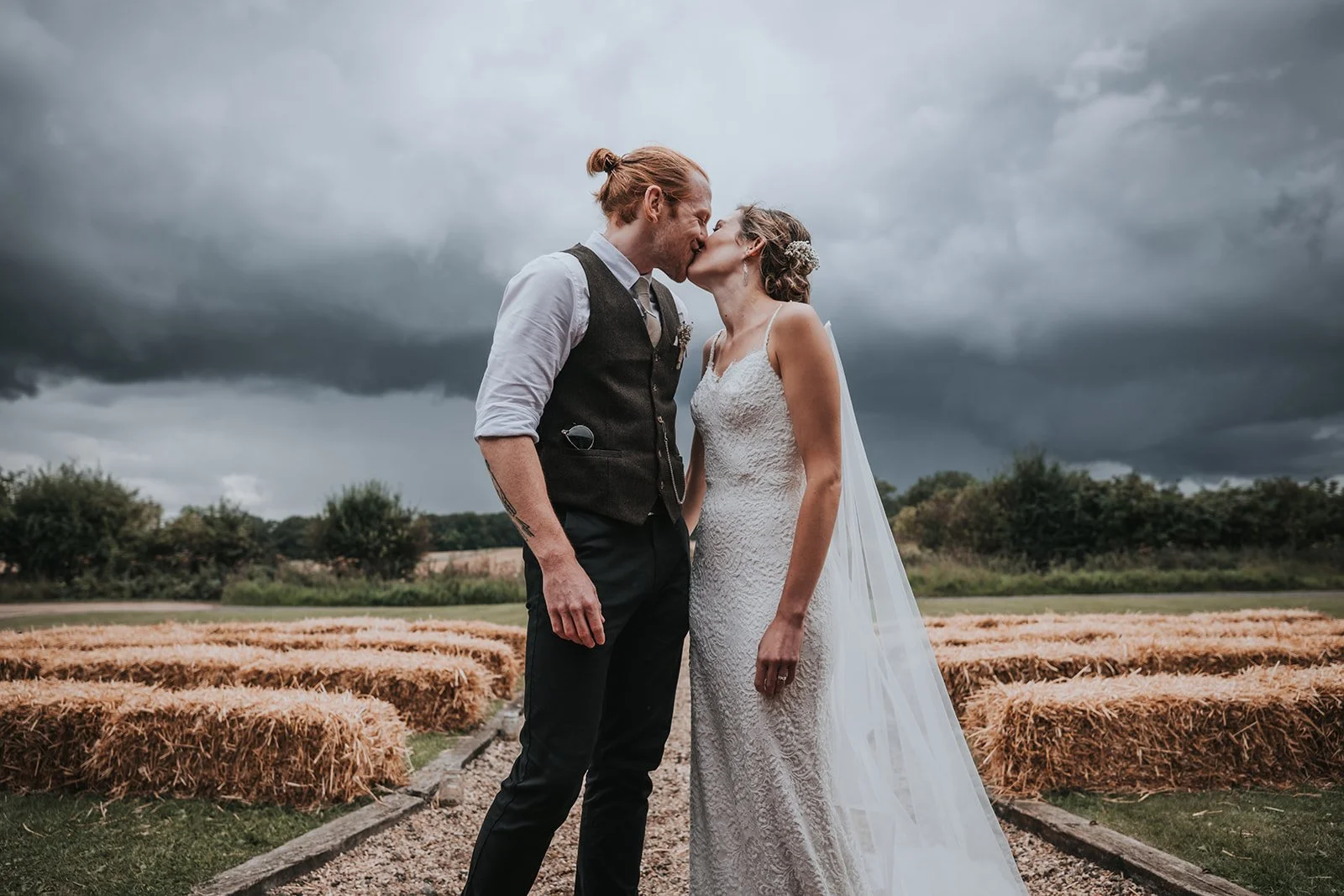 A bride and groom kiss outdoors in a field with hay bales and dark storm clouds in the sky.