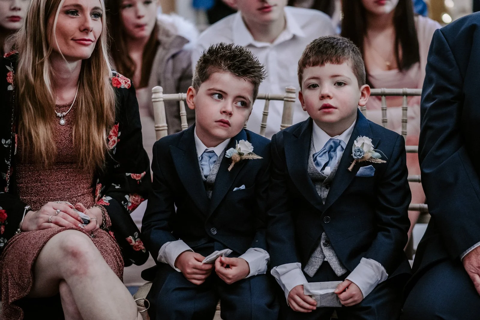 Two young boys dressed in formal suits sitting next to a woman at a wedding, with other guests in the background.