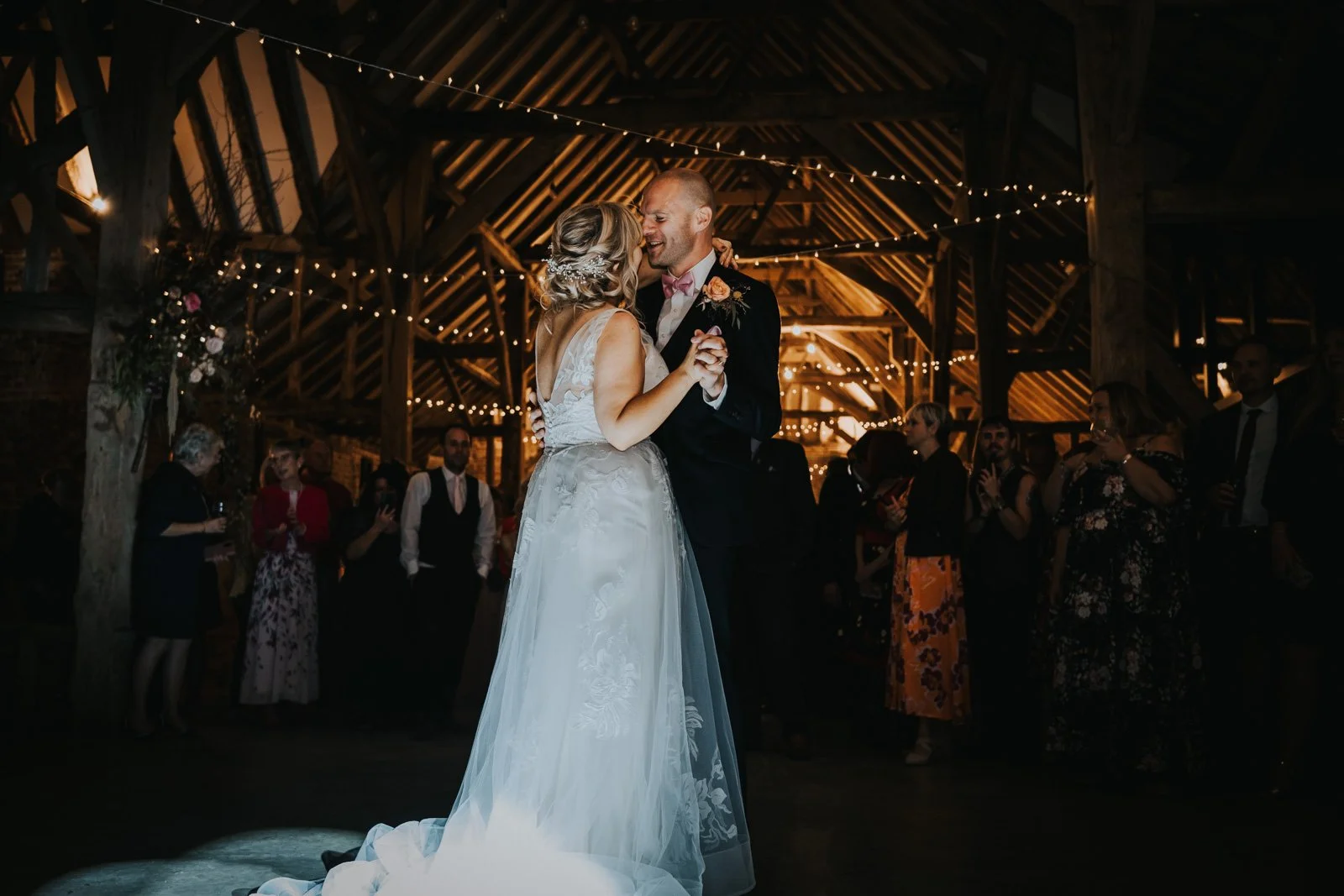 A bride and groom share their first dance at a wedding reception in a rustic barn decorated with string lights, with guests watching and celebrating.