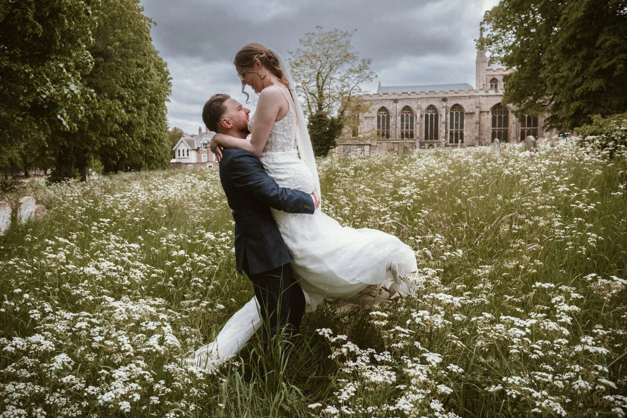 A couple in wedding attire, with the groom lifting the bride, standing amidst a field of white flowers in the Great Churchyard, Bury St Edmunds..