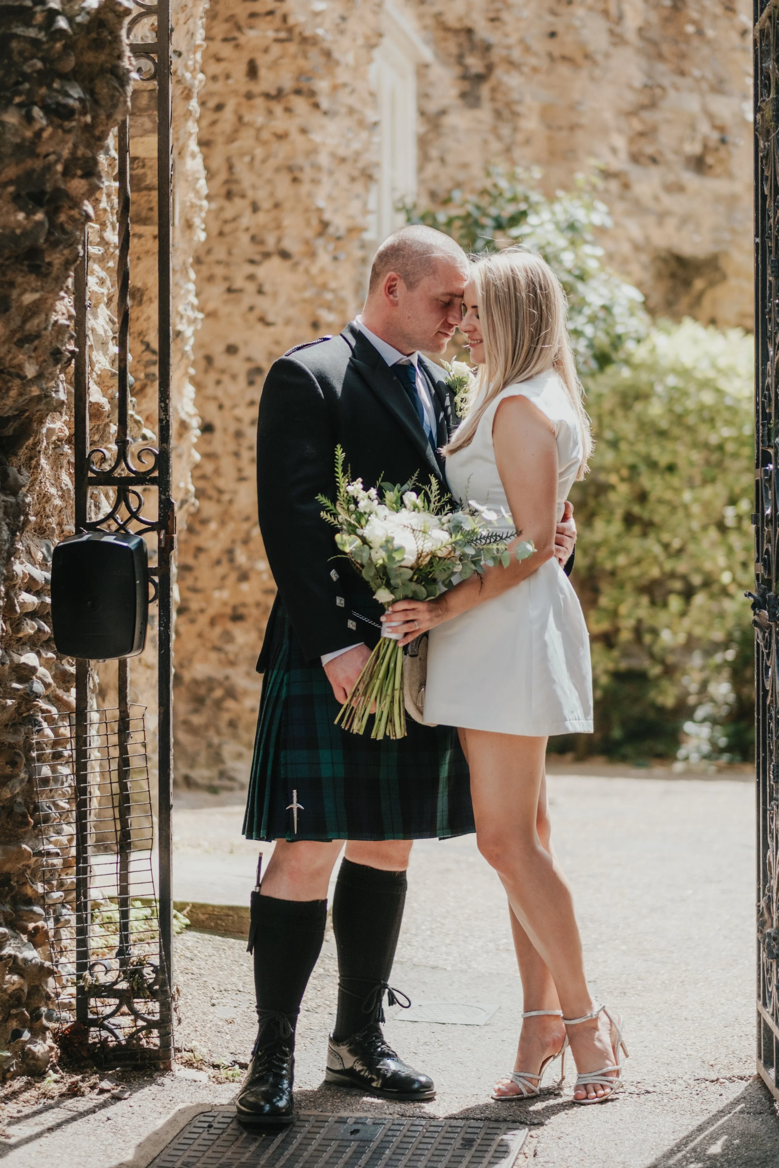 A groom dressed in a traditional Scottish kilt, jacket, and tie with a bride in a short white wedding dress are standing close together outdoors. The bride holds a bouquet of flowers. They touch foreheads and smile gently. 