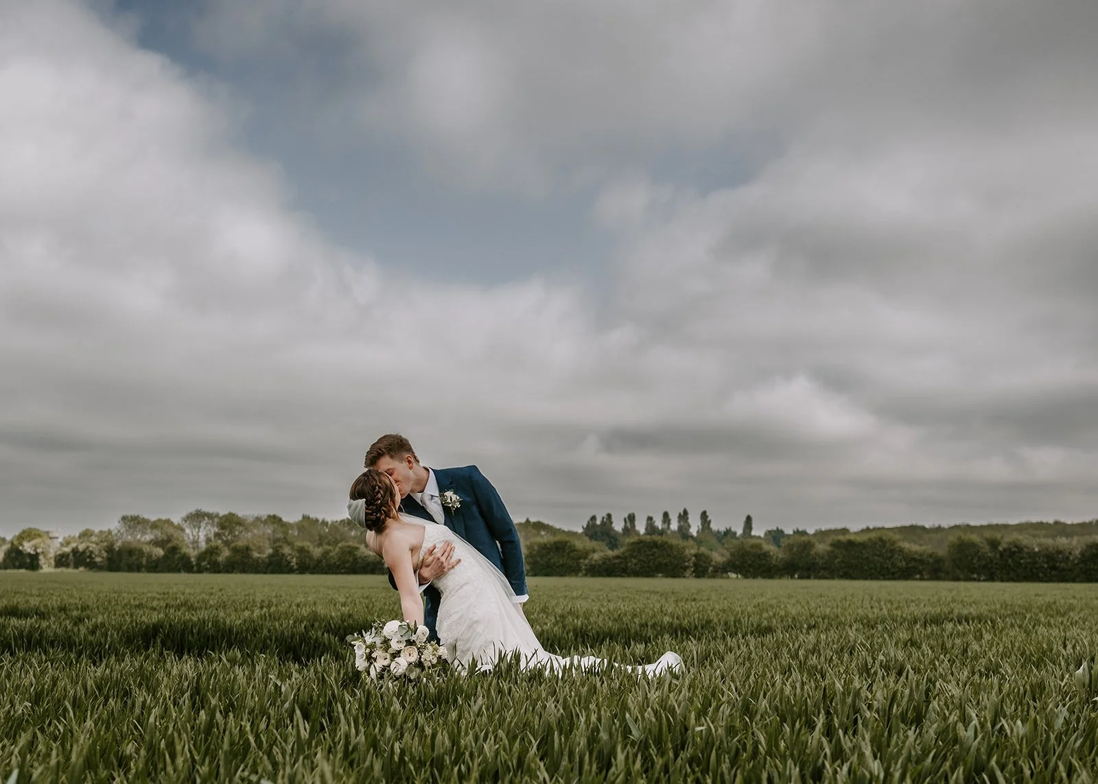 A bride and groom kissing in a grassy field on a cloudy day, with the bride holding a bouquet of flowers.
