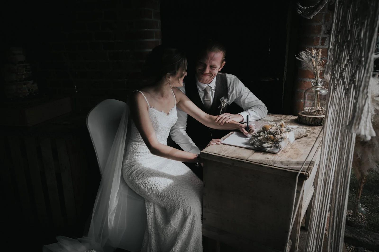 A bride and groom sitting together at a table, with the bride signing a document, in a cozy indoor setting with dim lighting, brick wall, and decorative dried flowers.