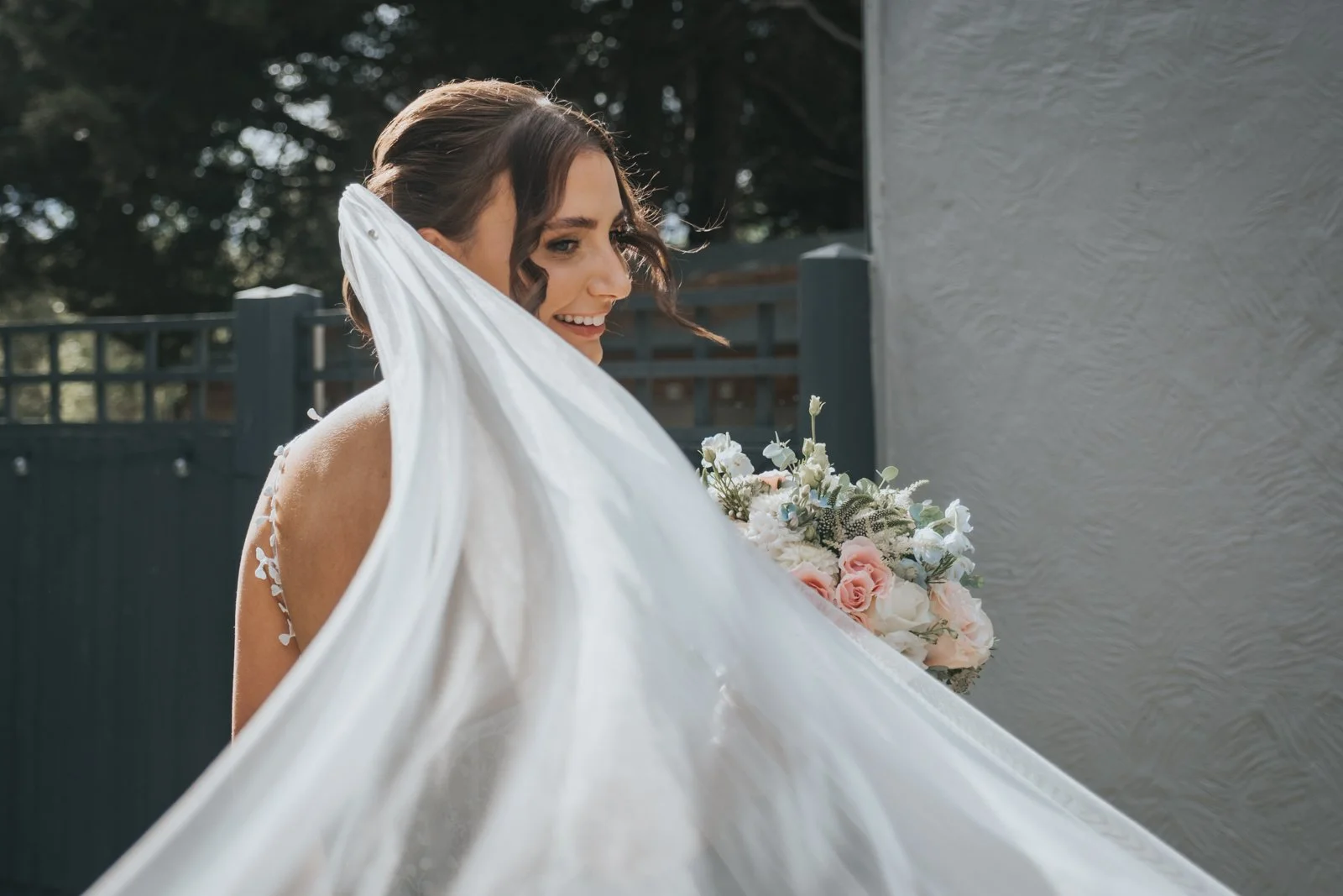 A bride holding a bouquet of pink and white flowers, smiling outdoors with greenery in the background.