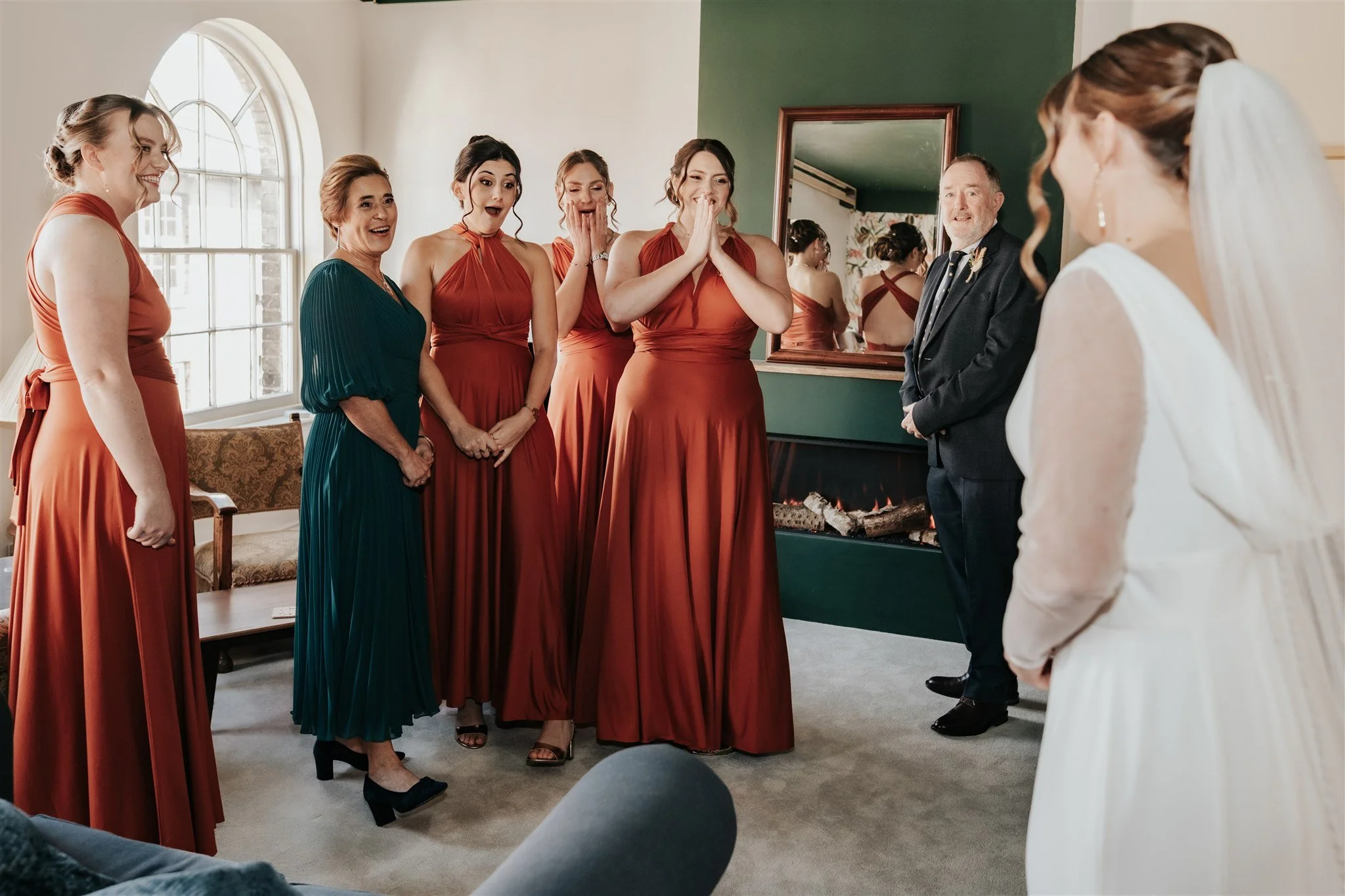 A group of women dressed in matching coral-colored bridesmaid dresses gathered in a room, reacting with surprise and joy to a bride in a white wedding gown. They are standing near a window with sunlight streaming in, and a man in a suit stands nearby