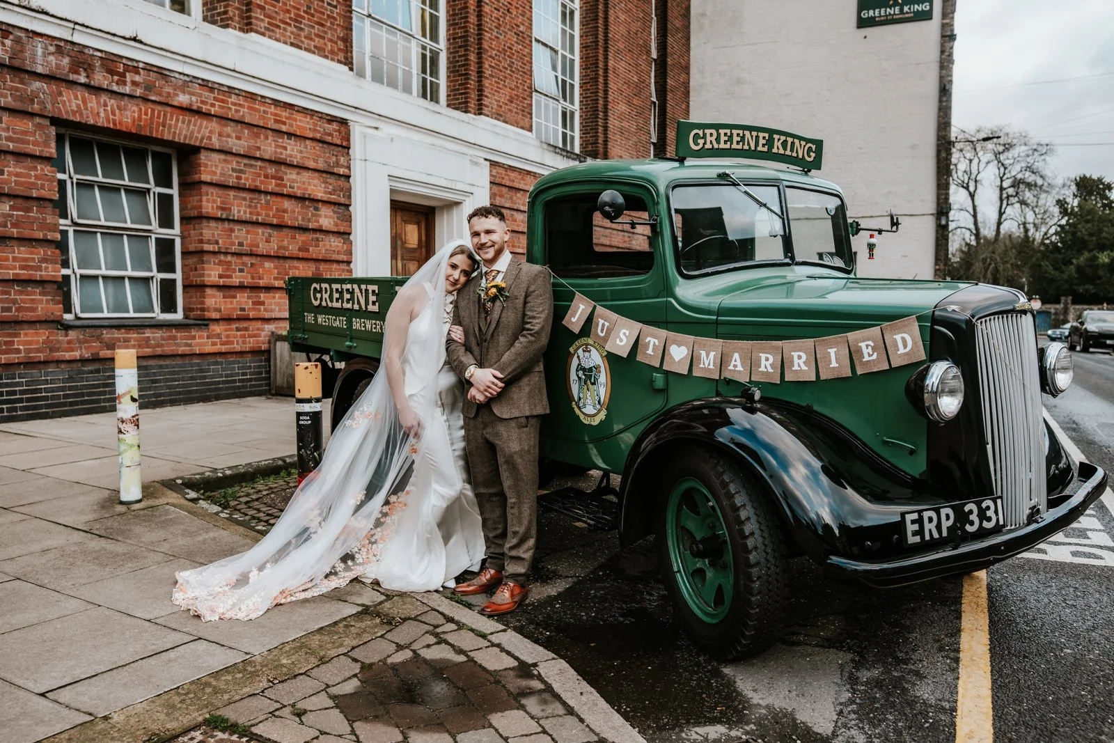 A newlywed couple in wedding attire standing next to a vintage green truck with a "Just Married" banner. The bride wears a white gown with floral details and a long veil. They hold hands outside the Greene King brewery, Bury St Edmunds.