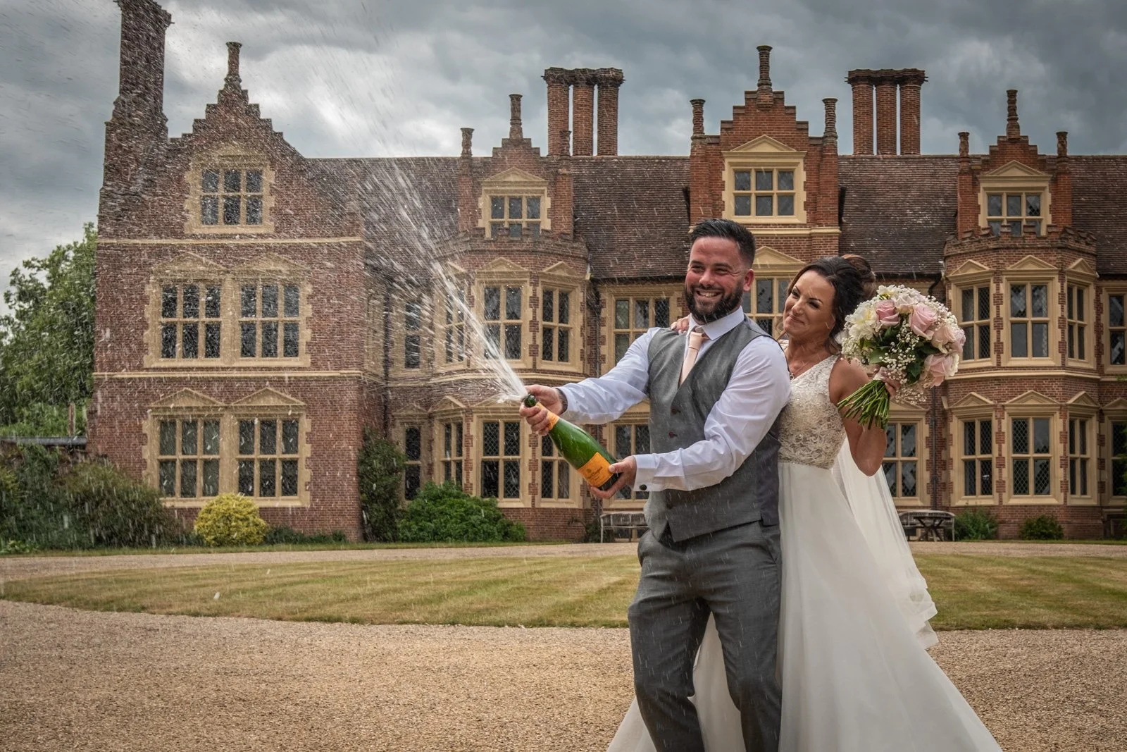 A newlywed couple celebrating outdoors in front of Haughley Park. The groom is smiling as he opens a bottle of champagne, causing it to spray, while the bride stands behind him holding a bouquet of pink and white flowers.