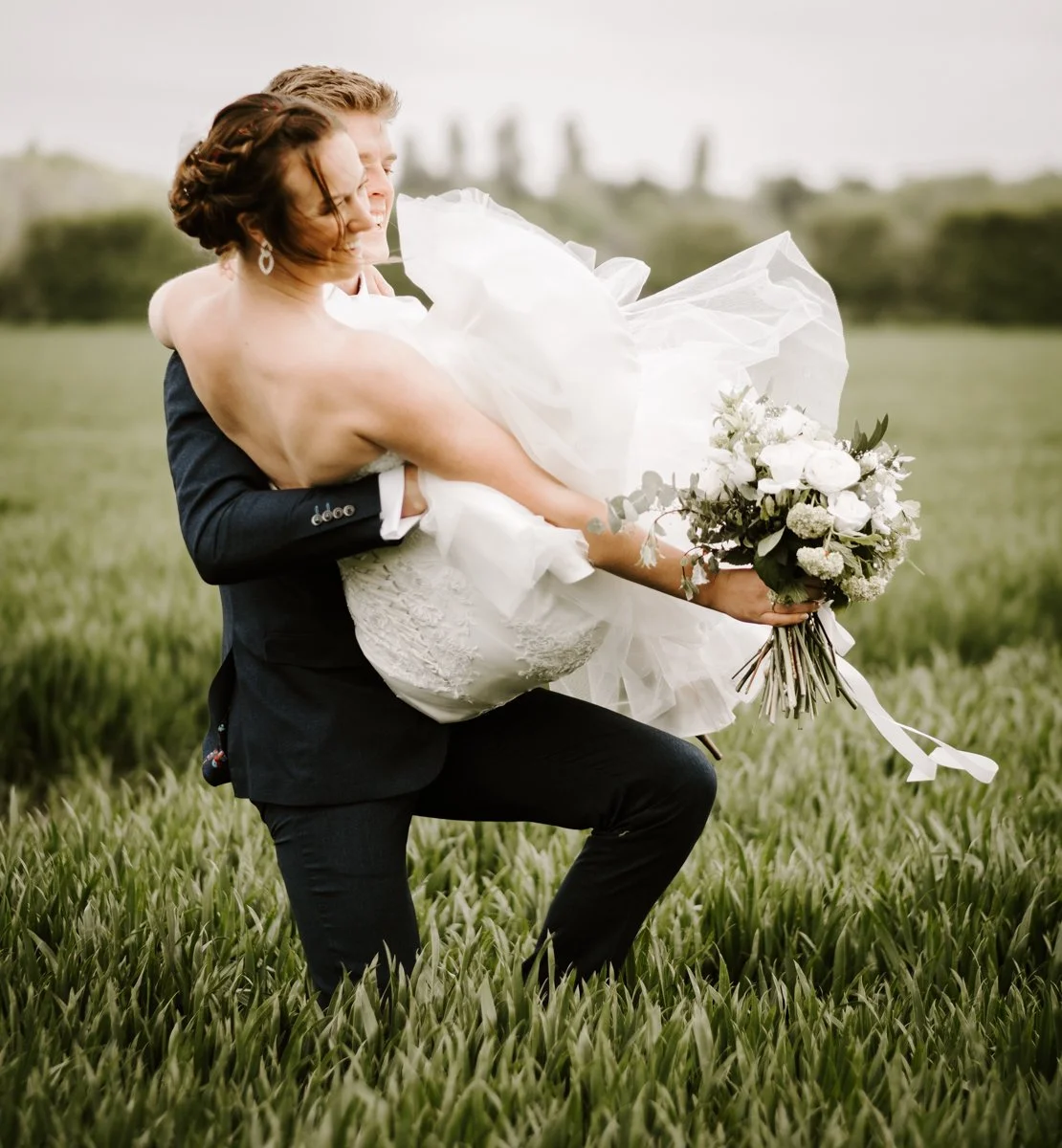 A groom lifting his bride in a grassy field, holding a bouquet of white and green flowers.