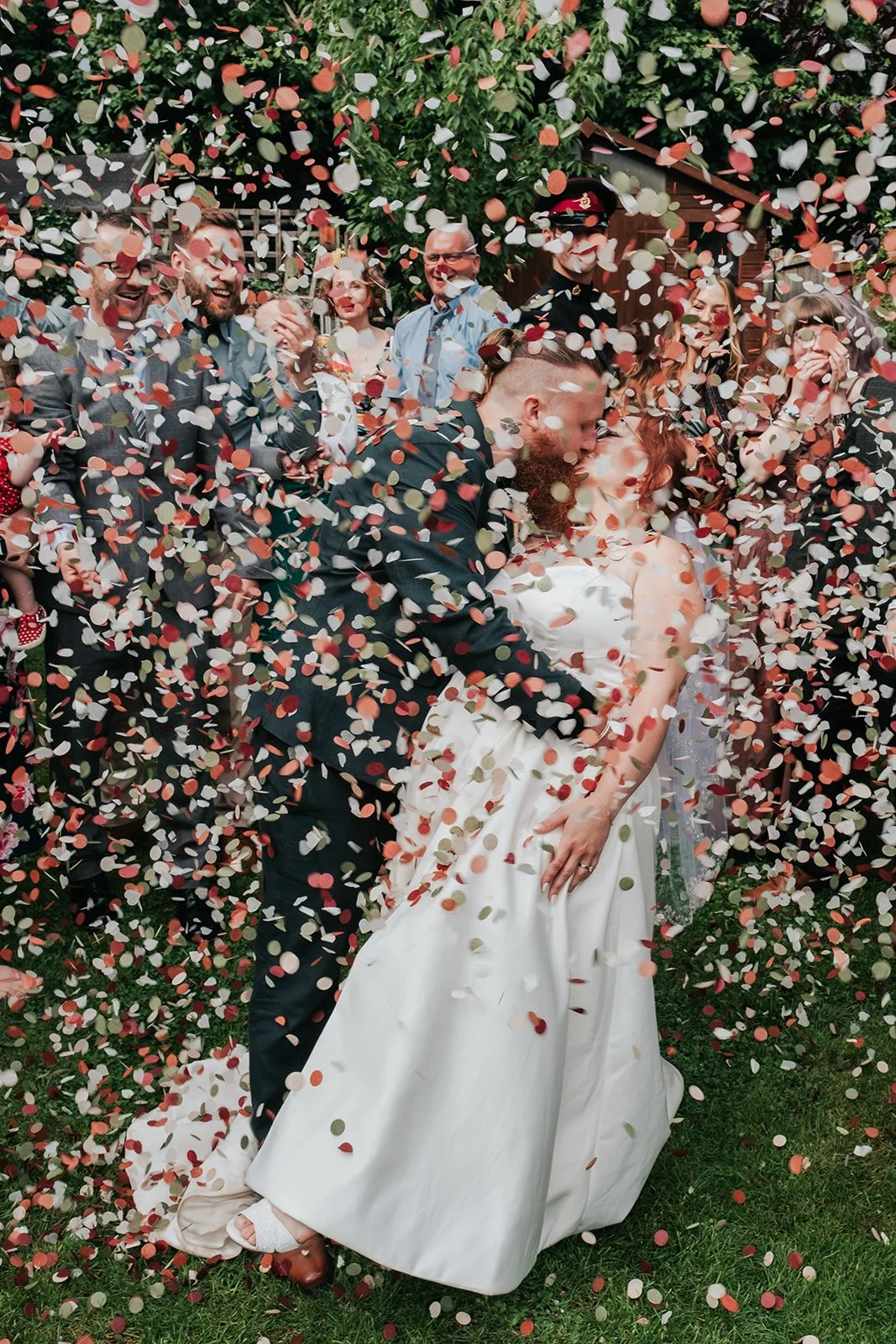 A wedding celebration with a bride and groom kissing under falling confetti, surrounded by friends and family outdoors.