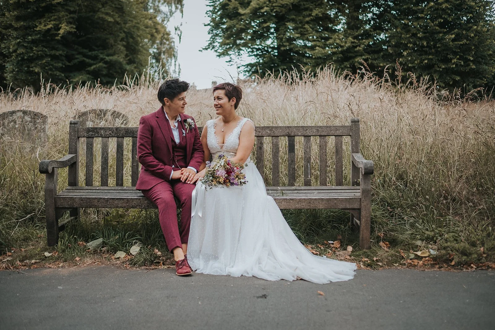 Two brides sitting on a wooden bench in a park, dressed in wedding attire, holding hands and smiling at each other, with tall grass and trees in the background.