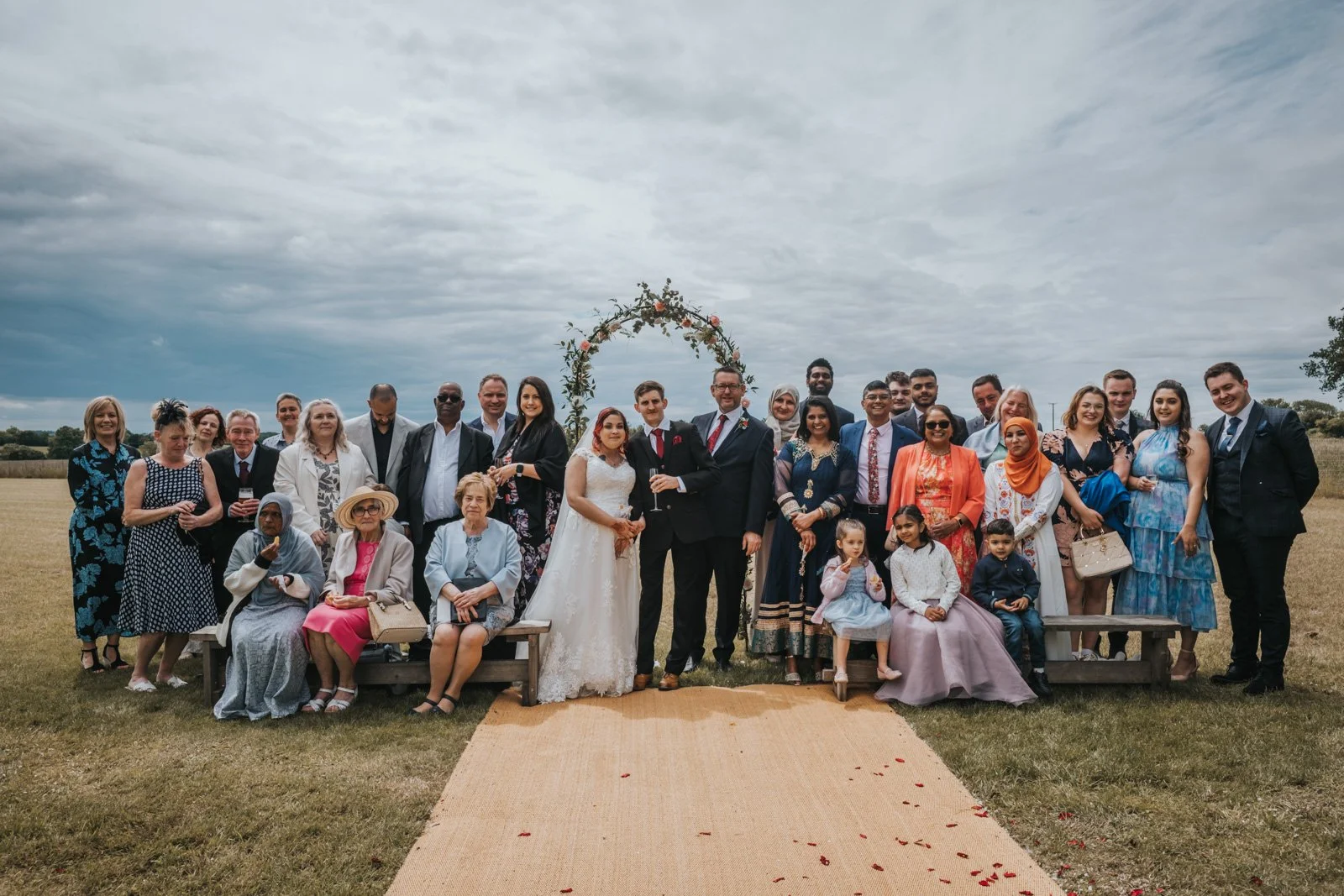 Group of people celebrating a wedding outdoors under a cloudy sky, with a bride and groom in the centre, family and friends surrounding them.