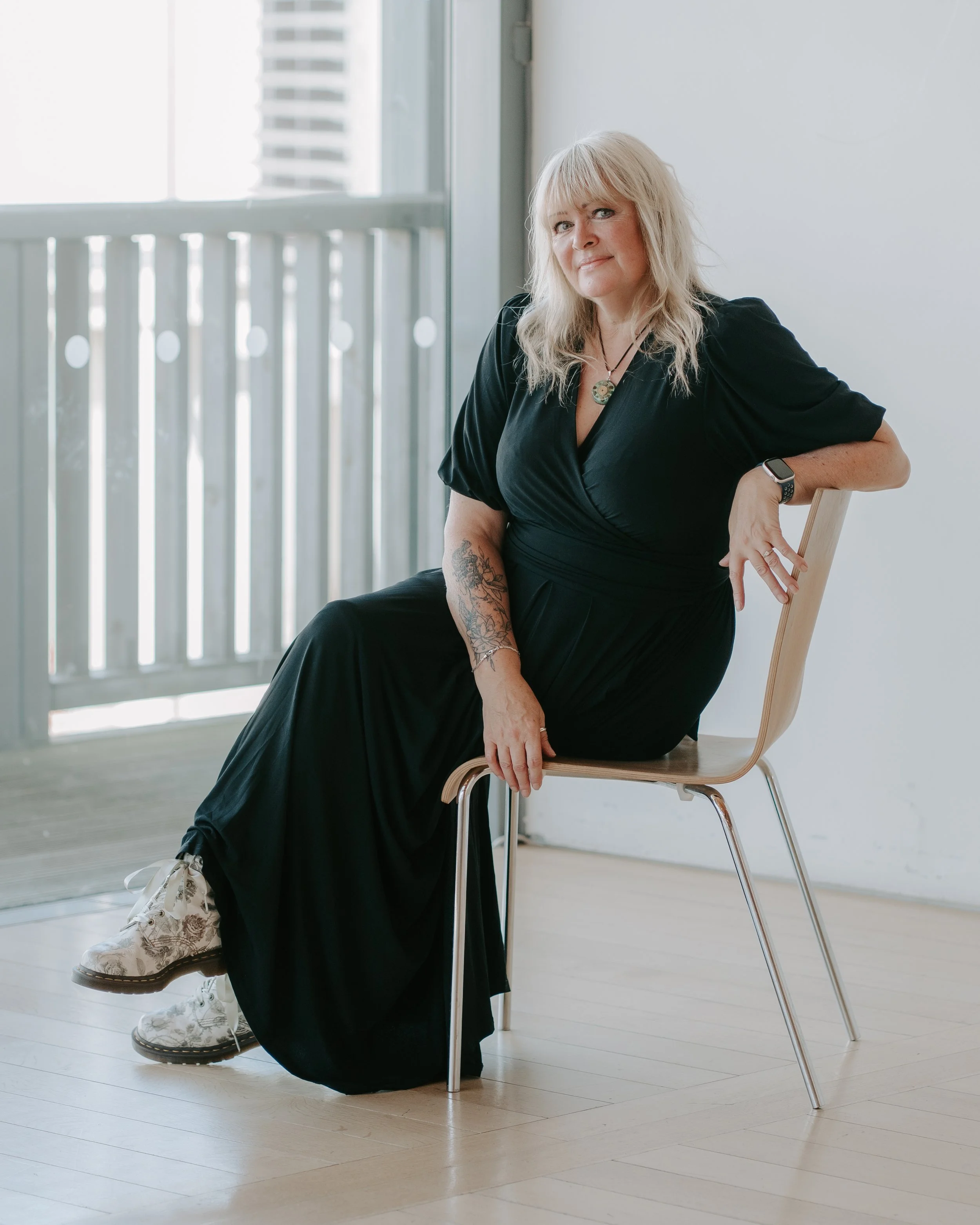 A woman with blonde hair, wearing a black dress, sitting on a wooden chair near a window with a white frame and railing, with a cityscape in the background. She has tattoos on her right forearm, is wearing a necklace, and a smartwatch on her left wrist.