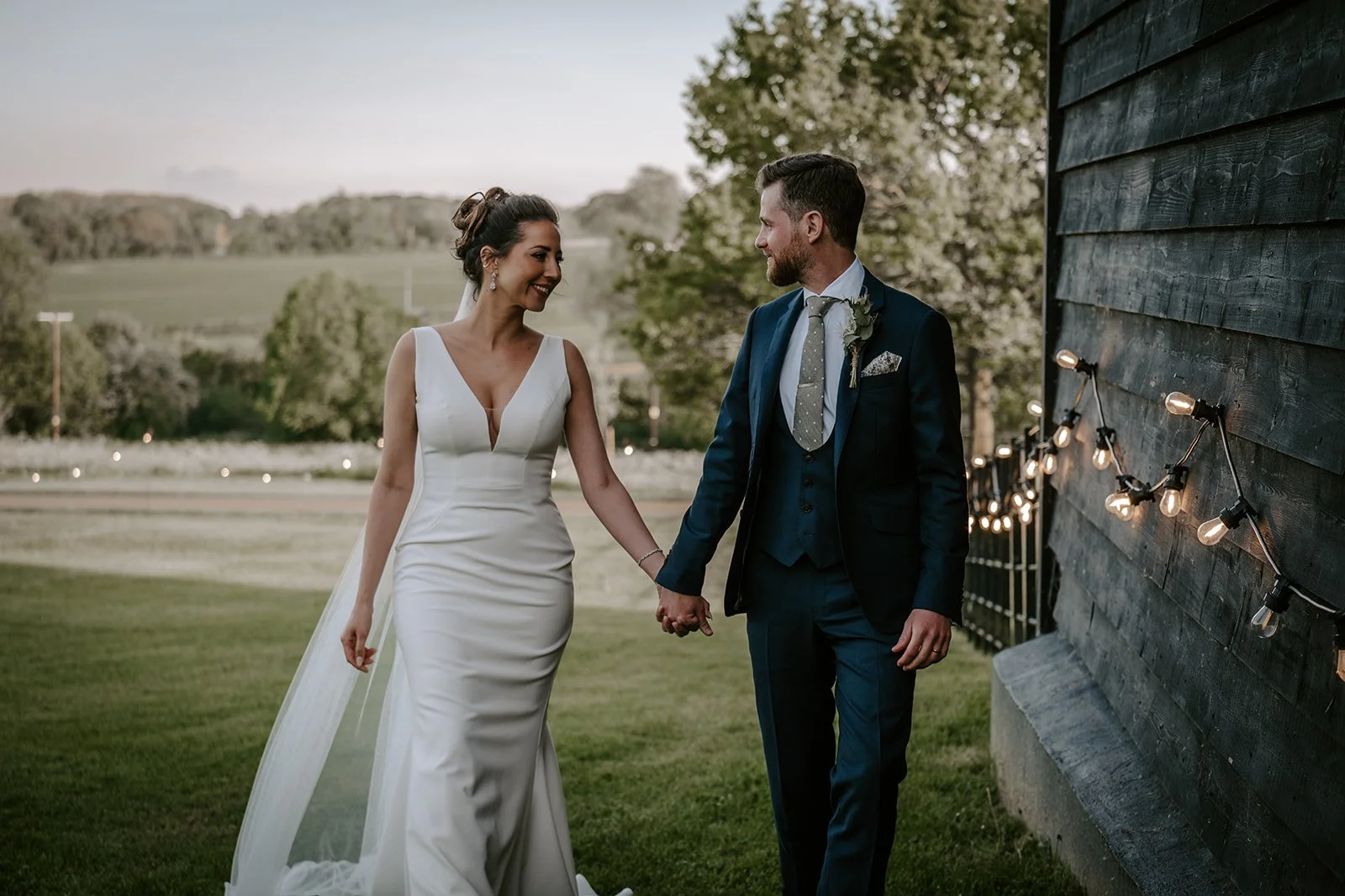Bride and groom holding hands outdoors during wedding, smiling at each other, with green landscape and string lights in the background at Suffolk Barn wedding venue..