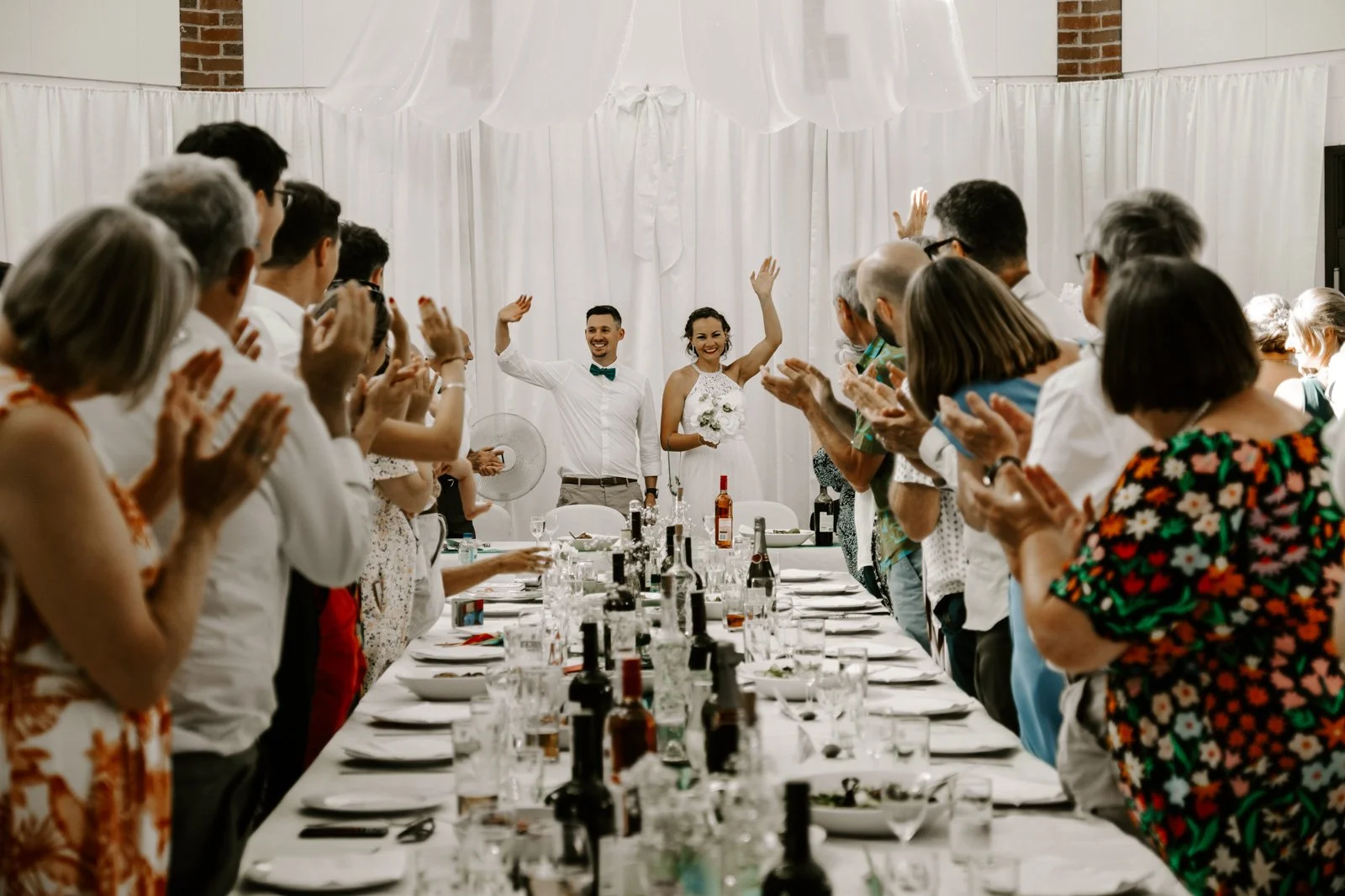 A newlywed couple standing at the end of a long banquet table, smiling and waving. The bride is holding a bouquet, and the groom has a green bow tie. Guests are clapping and standing around the table, which is set with wine bottles, glasses, and plat
