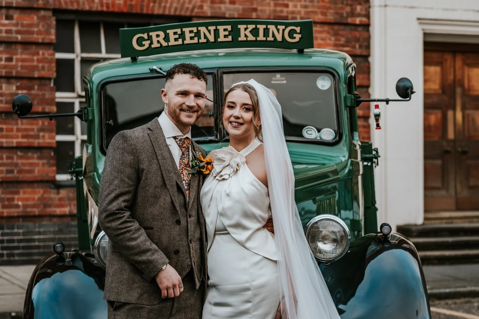A bride and groom smiling and standing in front of a vintage green dray with a sign that reads 'Greene King' on top. The groom is wearing a brown tweed suit and the bride is wearing a white wedding dress with a veil.