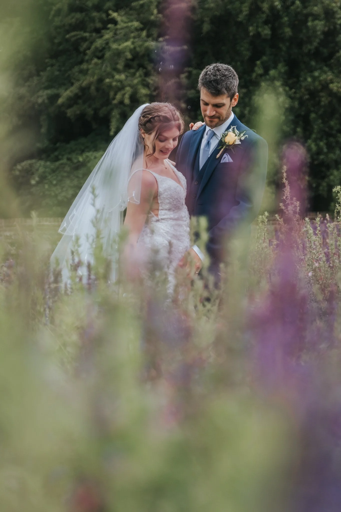 A bride and groom standing close together outdoors, surrounded by green foliage and purple flowers, at Abbey Gardens, Bury St Edmunds.